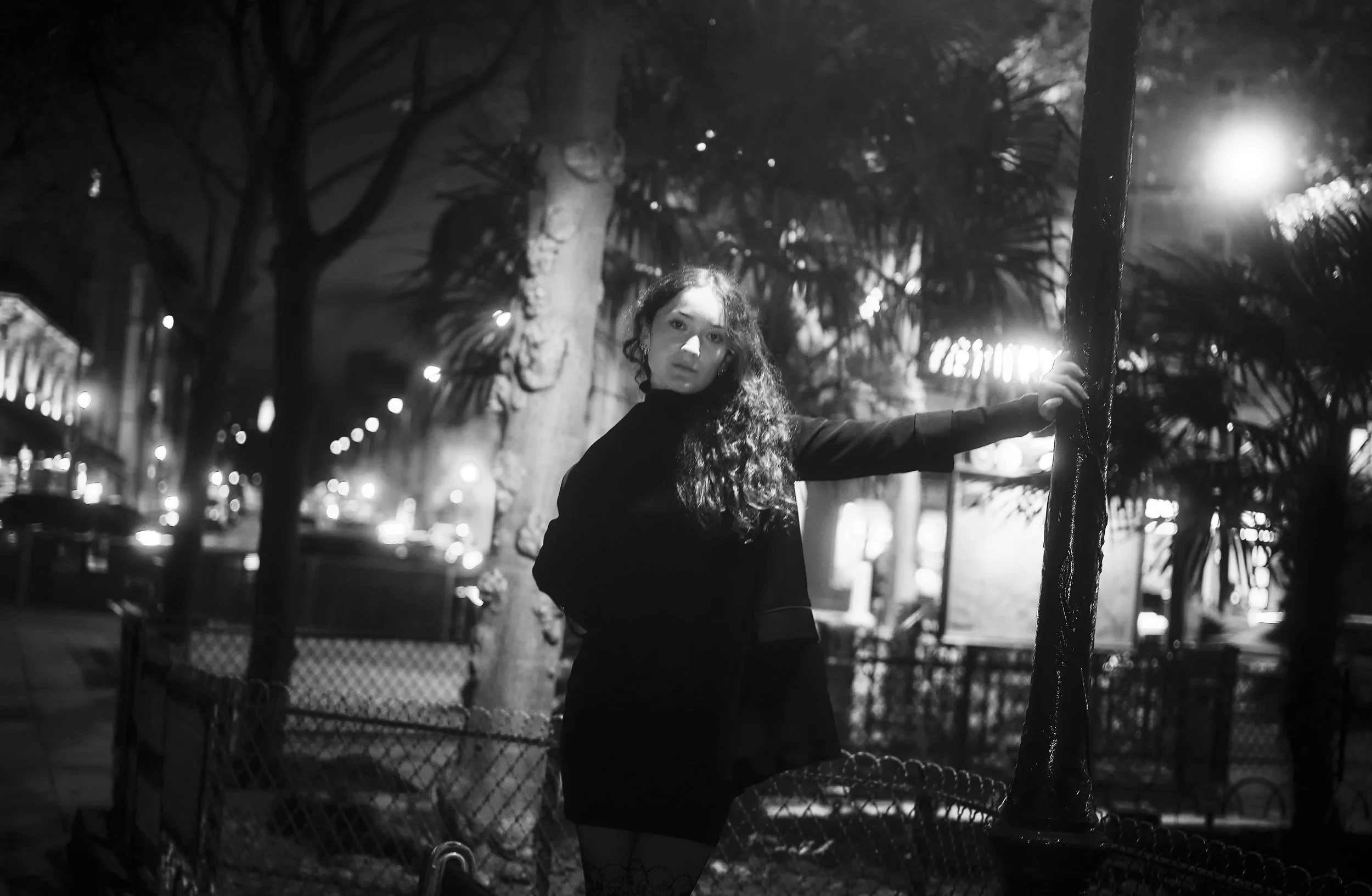 Photo pour séance Portrait Lifestyle. Femme debout près d'un lampadaire dans une rue de nuit, entourée d'arbres et de lampadaires, en noir et blanc.