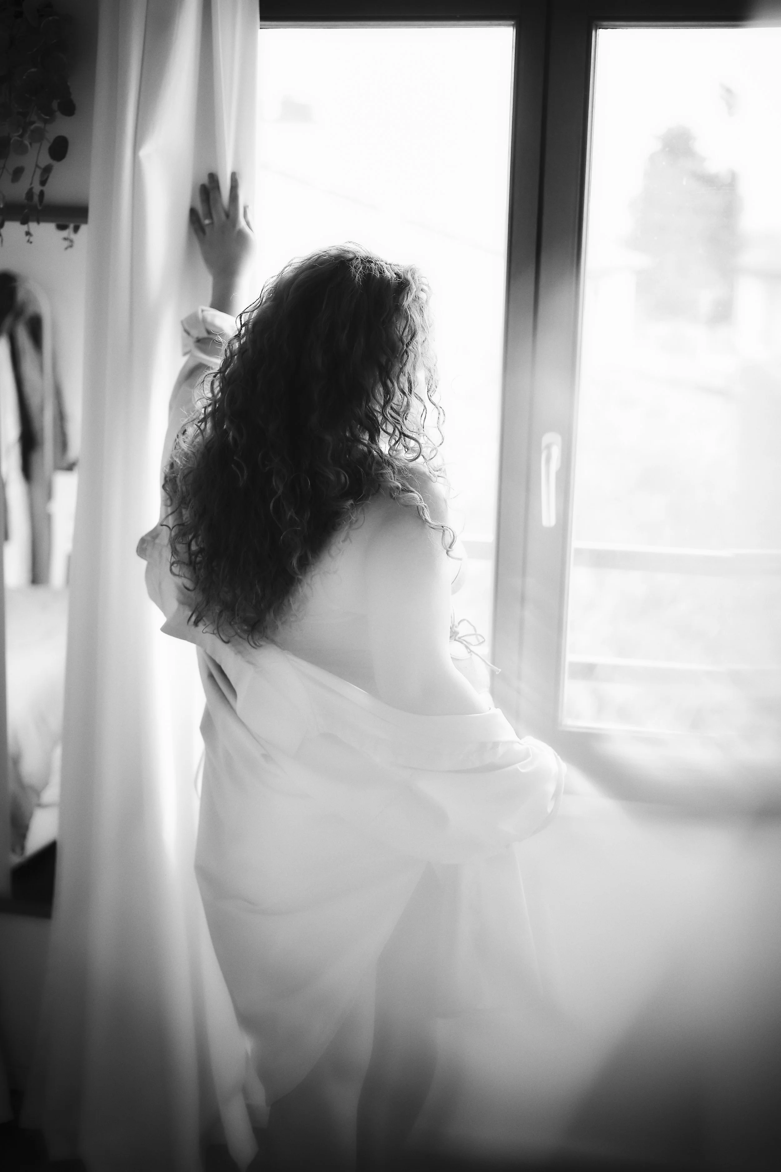 Photo pour séance Portrait intimiste Anonyme. Une femme aux cheveux bouclés, vue de dos, regarde par une grande fenêtre dans une pièce lumineuse.