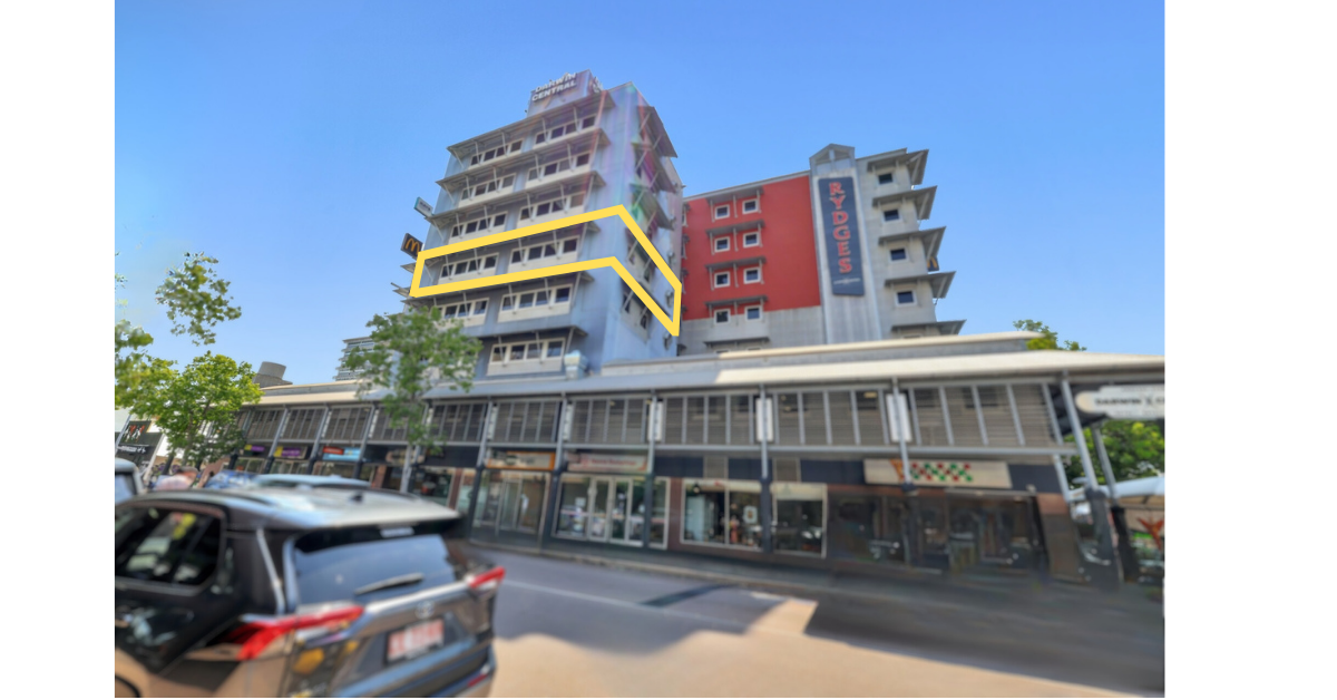 A multi-story building with retail shops at the ground level and apartments or hotel rooms above, with a parking lot and cars in front, and blue sky overhead.