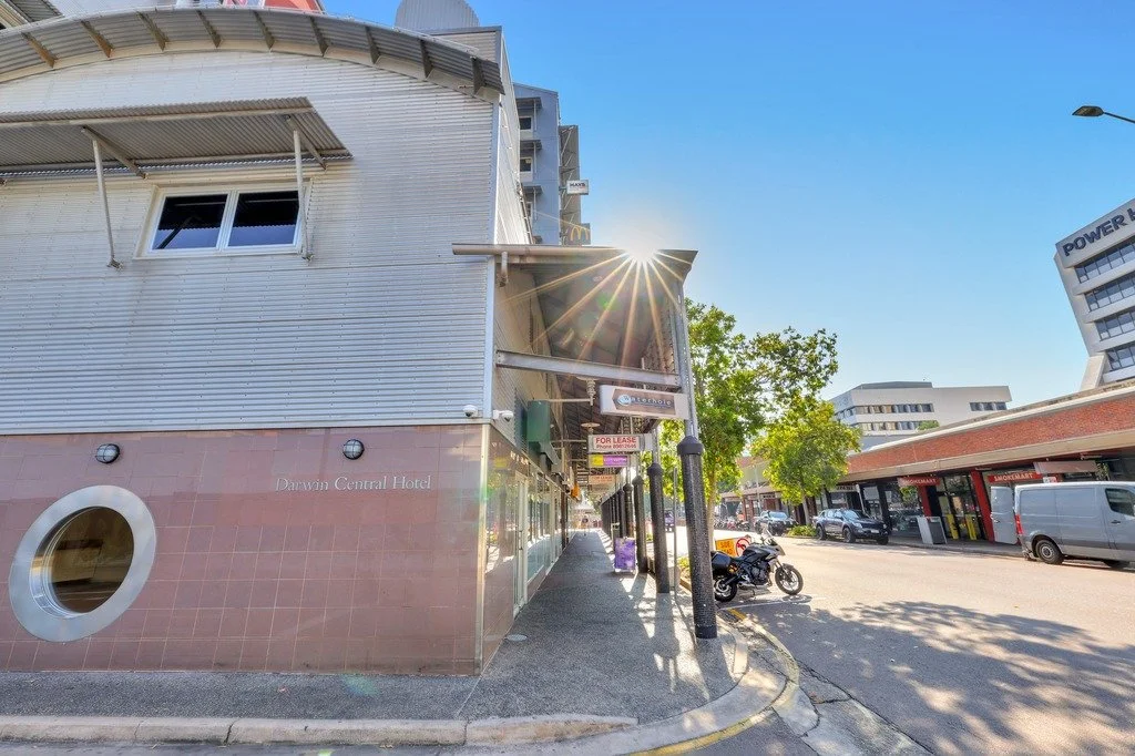 Street view with a modern hotel building on the left, sidewalk, parked motorcycle, trees, and other buildings in the distance on a sunny day with clear blue sky.