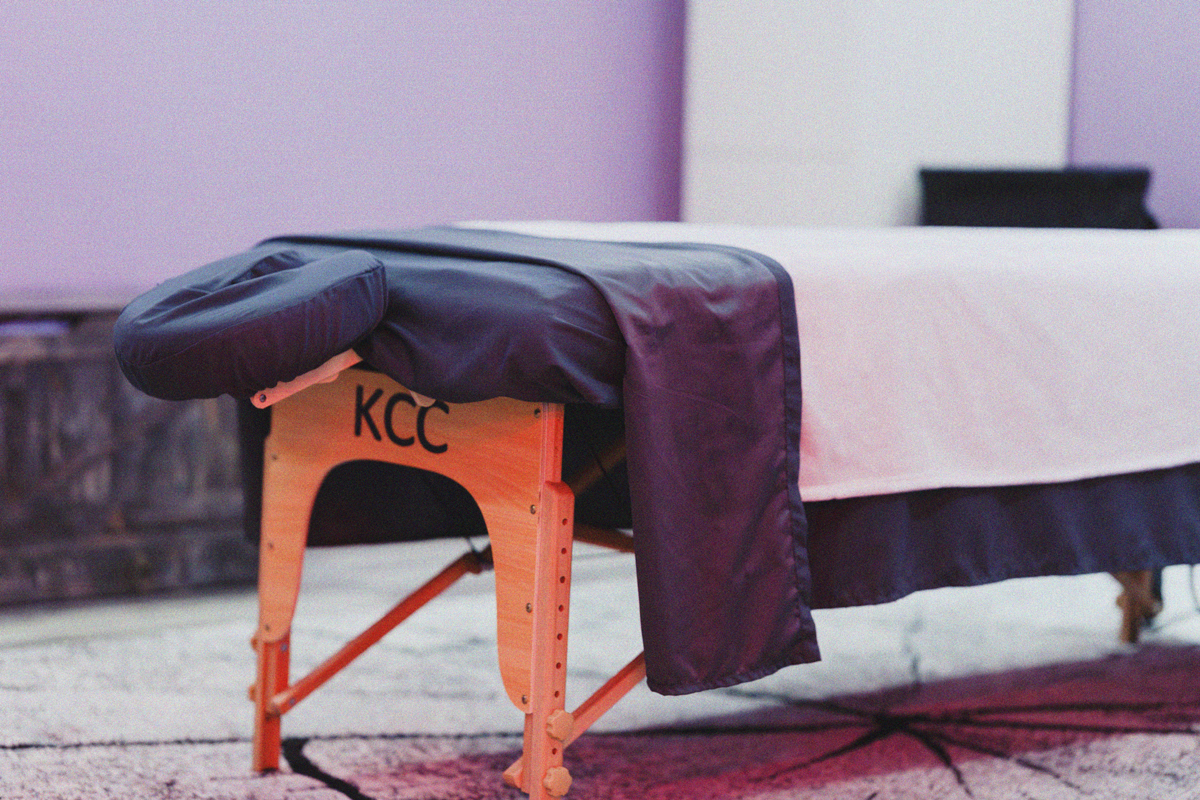 Massage table with a black cover and a purple sheet, situated on a patterned rug with a stone wall in the background.