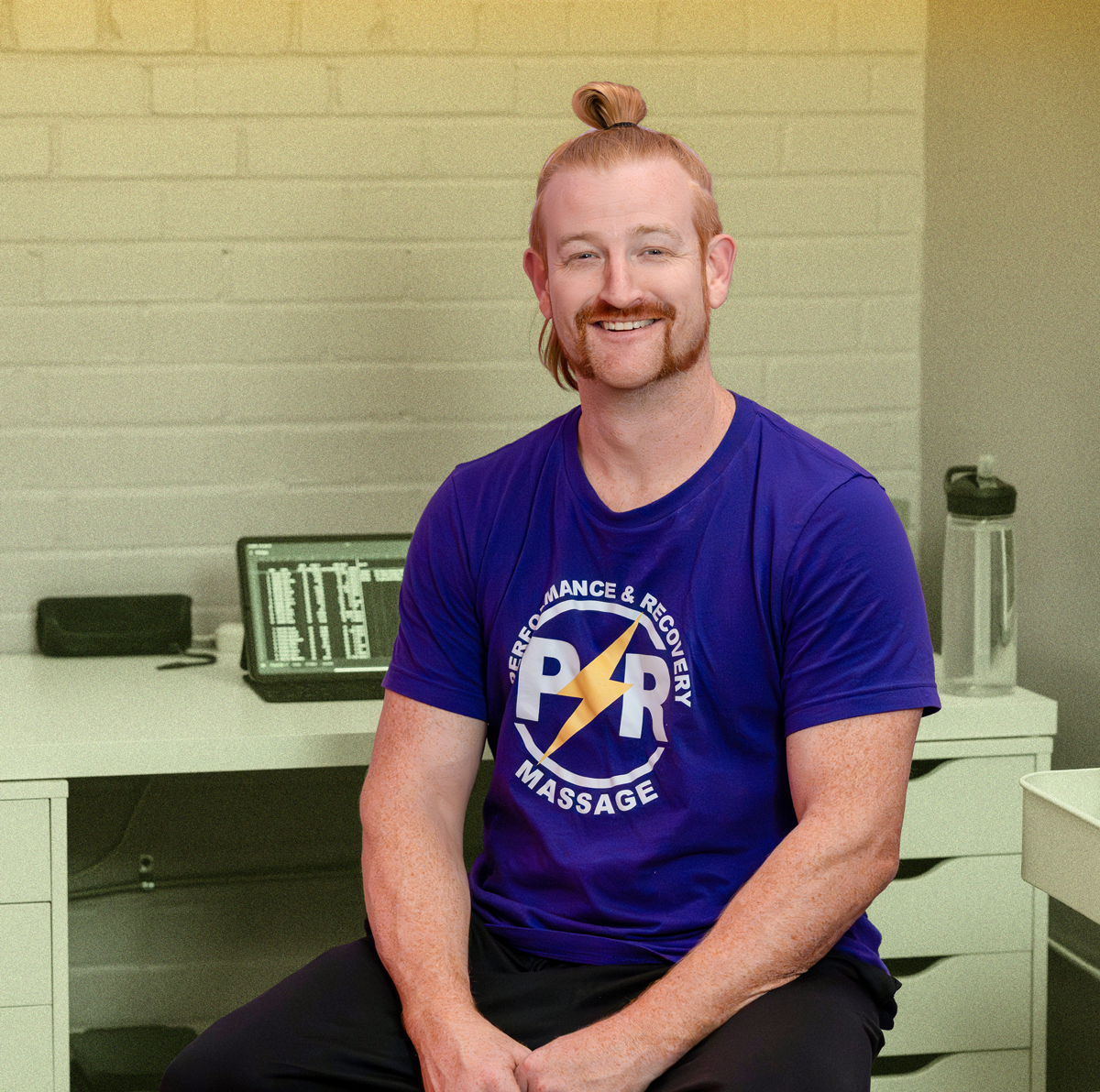A man with red hair styled in a top bun, wearing a blue T-shirt with a massage logo, sitting in an office.