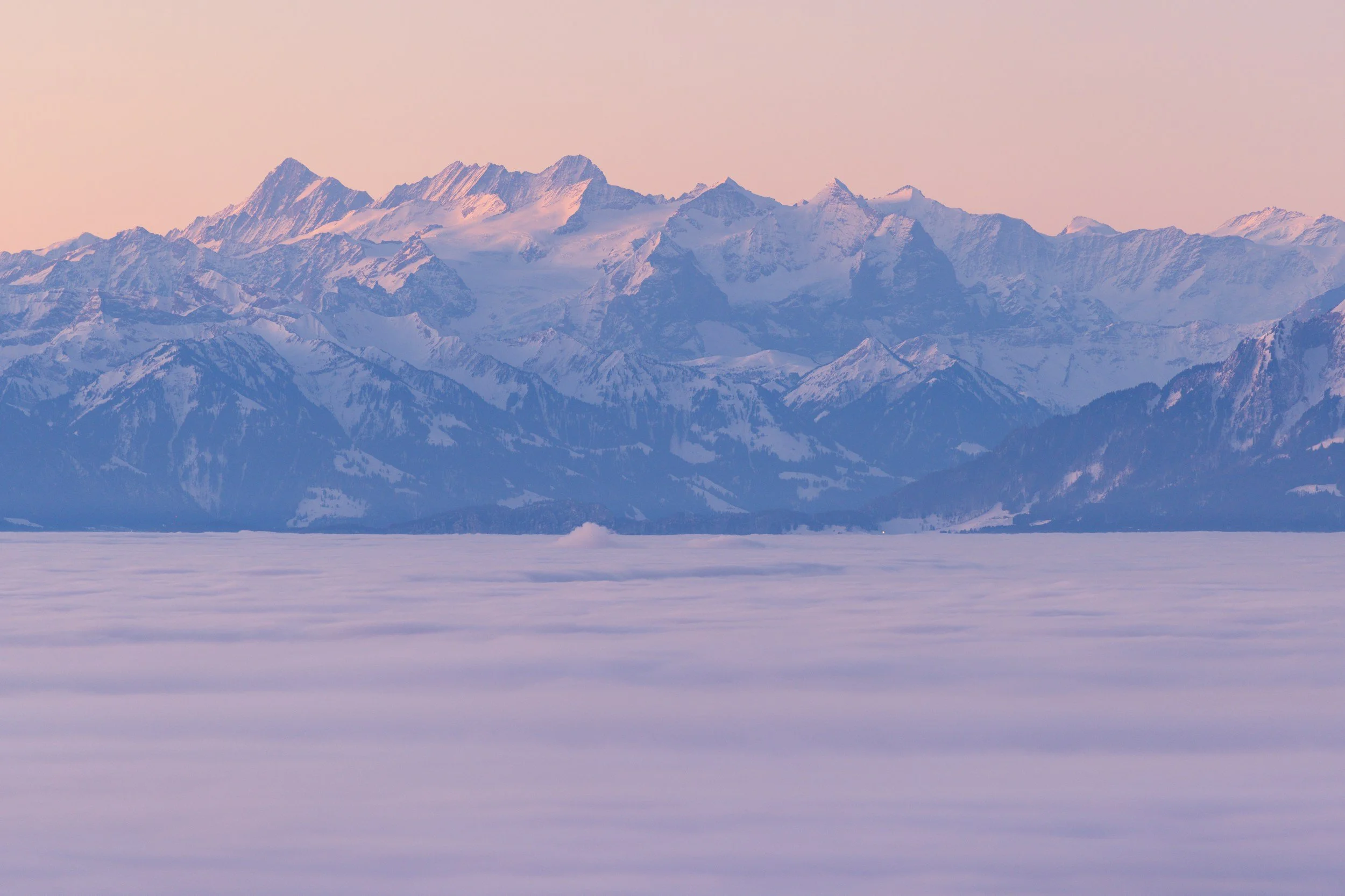 Blick auf verschneite Gebirgskette bei Sonnenaufgang mit rosanem Himmel, im Vordergrund eine schneebedeckte Ebenen