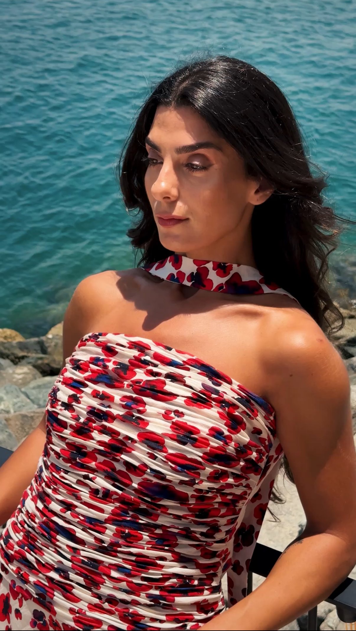 A woman with dark wavy hair wearing a sleeveless red, white, and blue floral dress sitting outdoors near a rocky shoreline with blue water in the background.