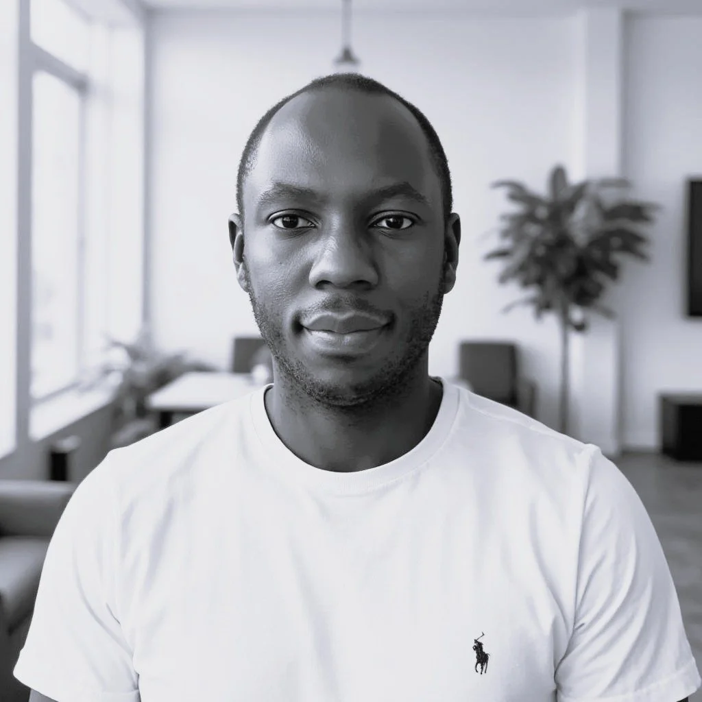 A young man with short hair and a slight smile, wearing a white Ralph Lauren Polo t-shirt, standing in a modern office or living space with large windows, a table, chairs, and a plant in the background.