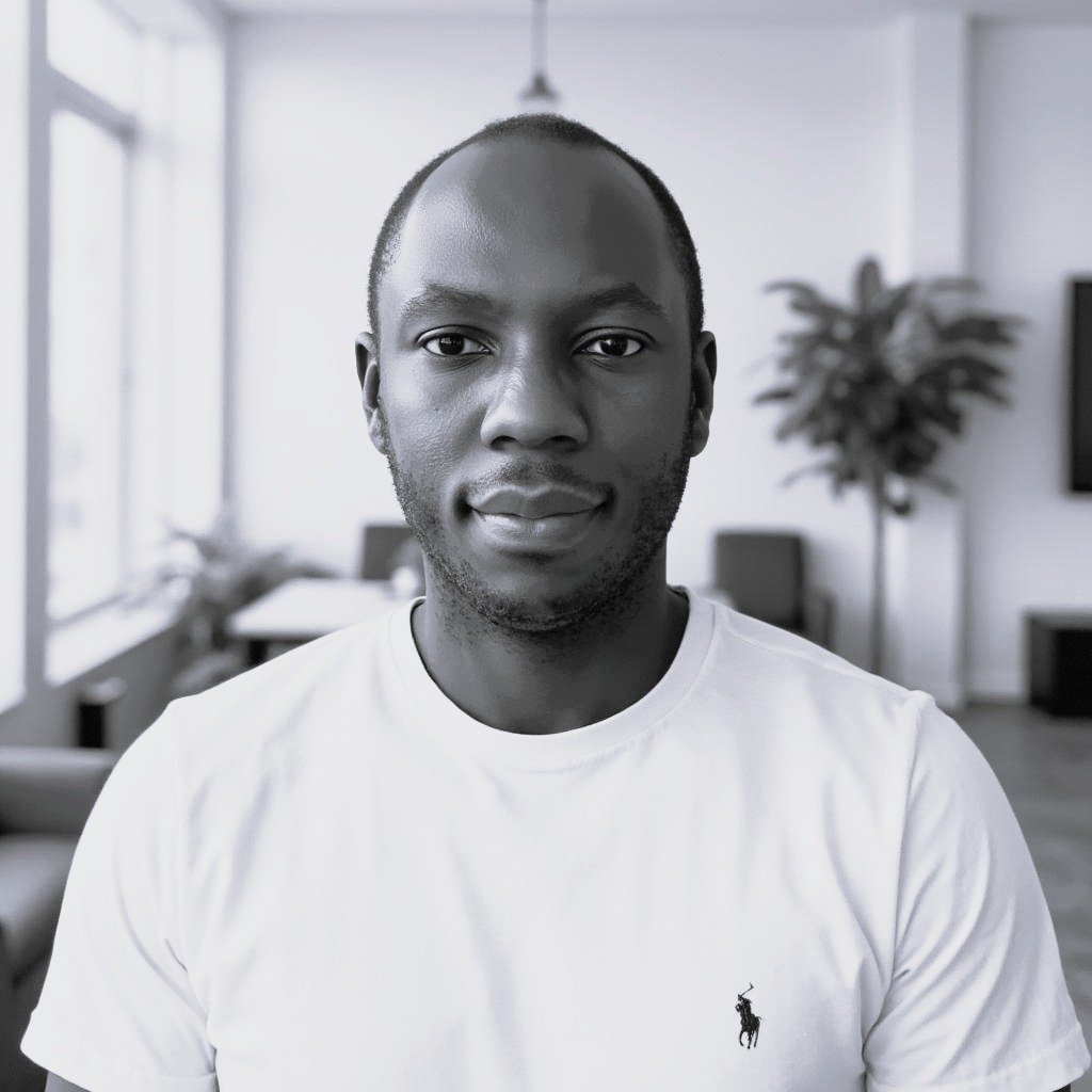A young man with a shaved head and a slight smile, wearing a white T-shirt, standing in a modern, well-lit living room with large windows, a TV, and a potted plant in the background.