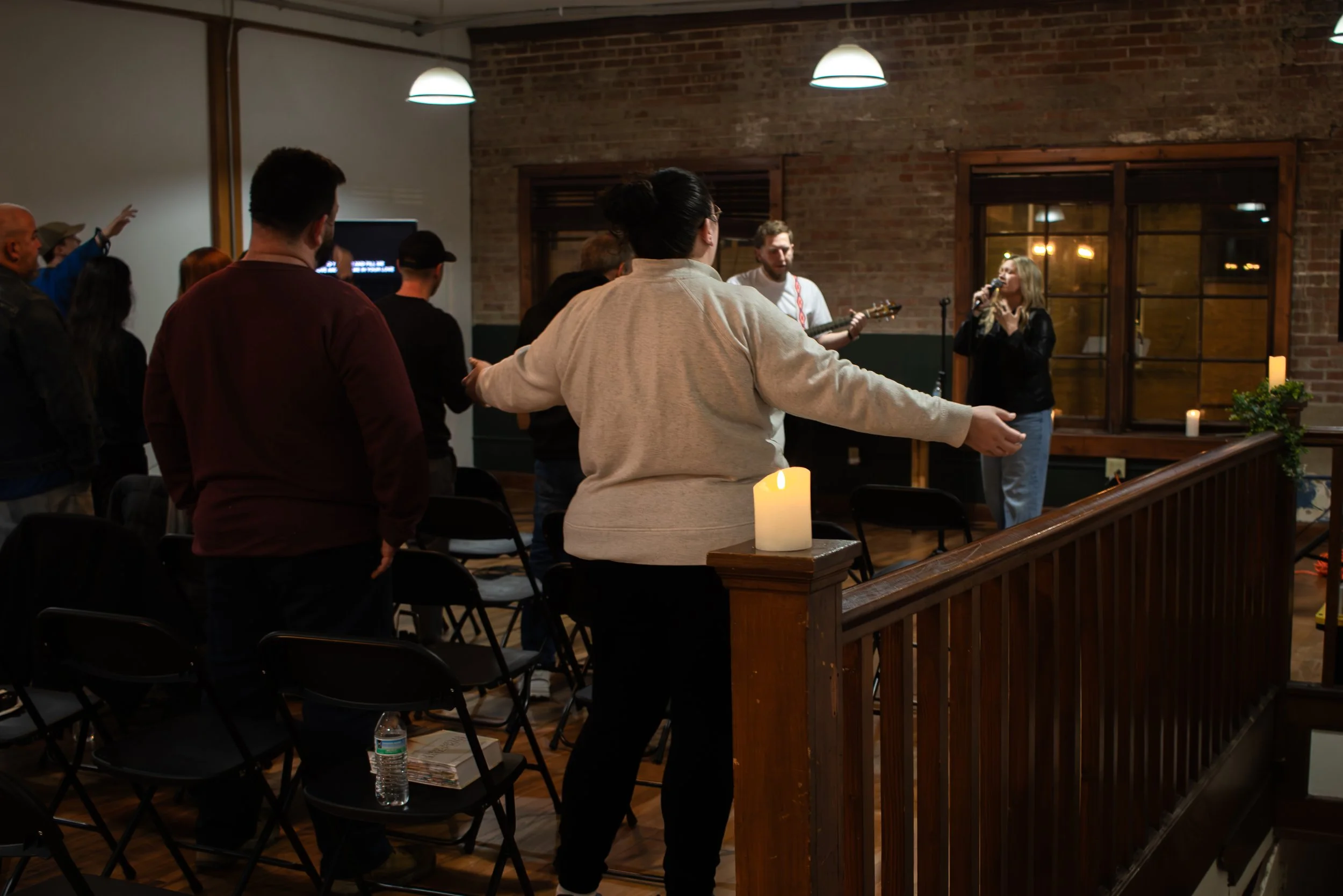 People in a cozy brick-walled room attending a music event with a singer and guitarist, some with arms raised, candles on a wooden ledge, chairs, and a water bottle on a chair.