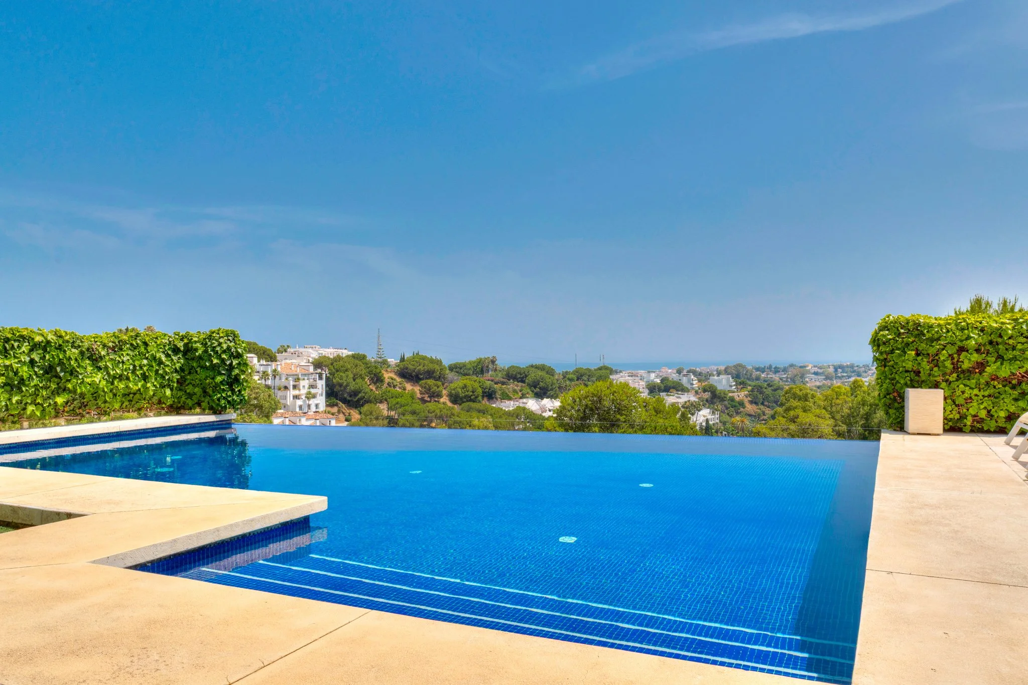 Infinity pool with blue water and beige pool deck, surrounded by green bushes and a clear blue sky in a scenic landscape