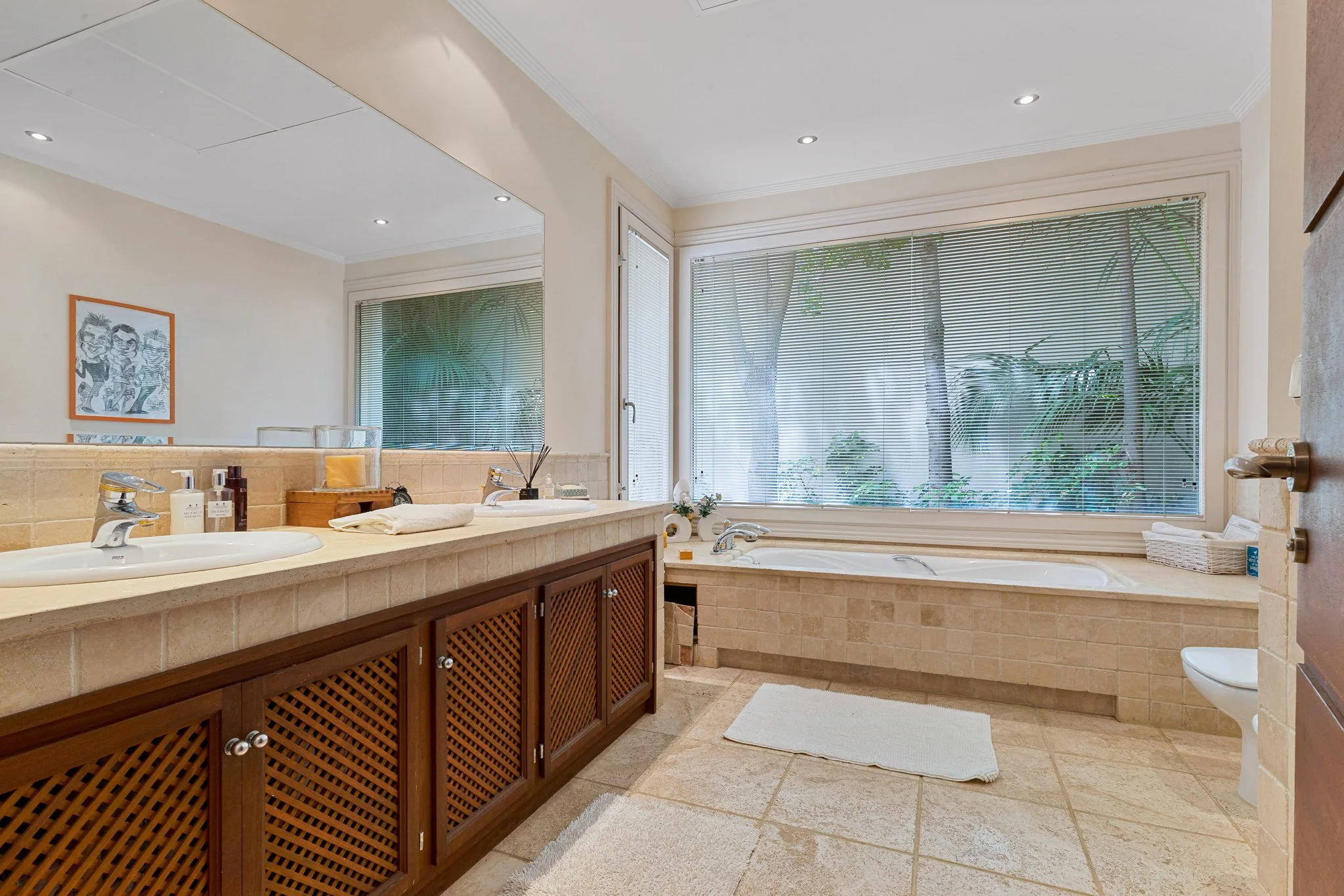 Bathroom with double sinks, a large mirror, a bathtub with a view of greenery through window blinds, and a small basket on the right side.
