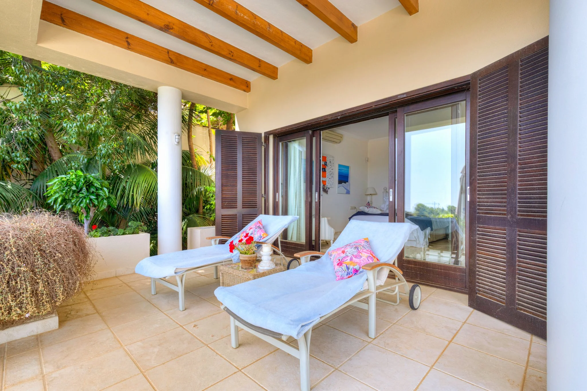 Balcony with two lounge chairs, a small table with a potted plant, overlooking greenery and a bedroom with sliding glass doors.