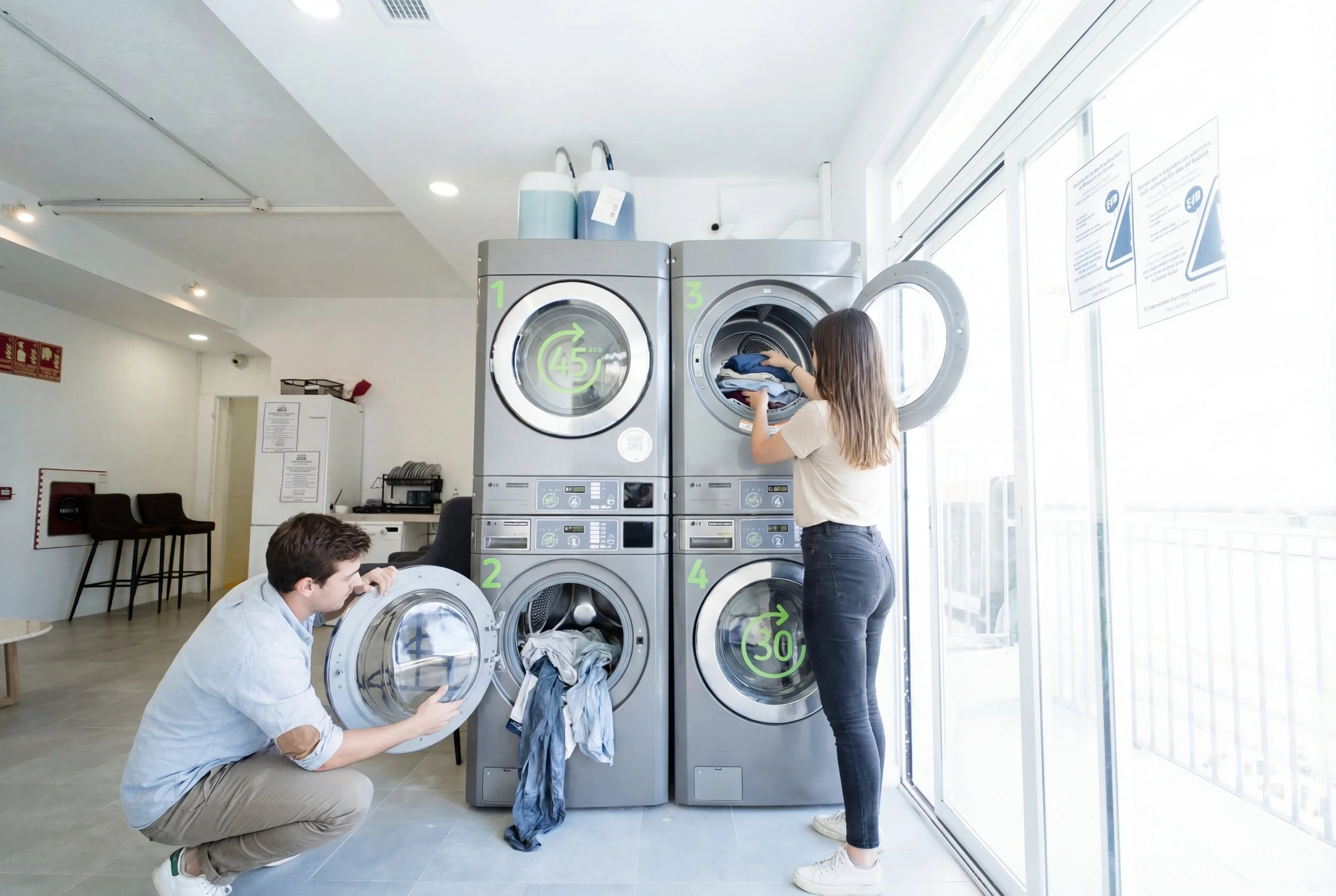 Lavandería con lavadoras y secadoras industriales; dos huéspedes cargan ropa junto a los ventanales.