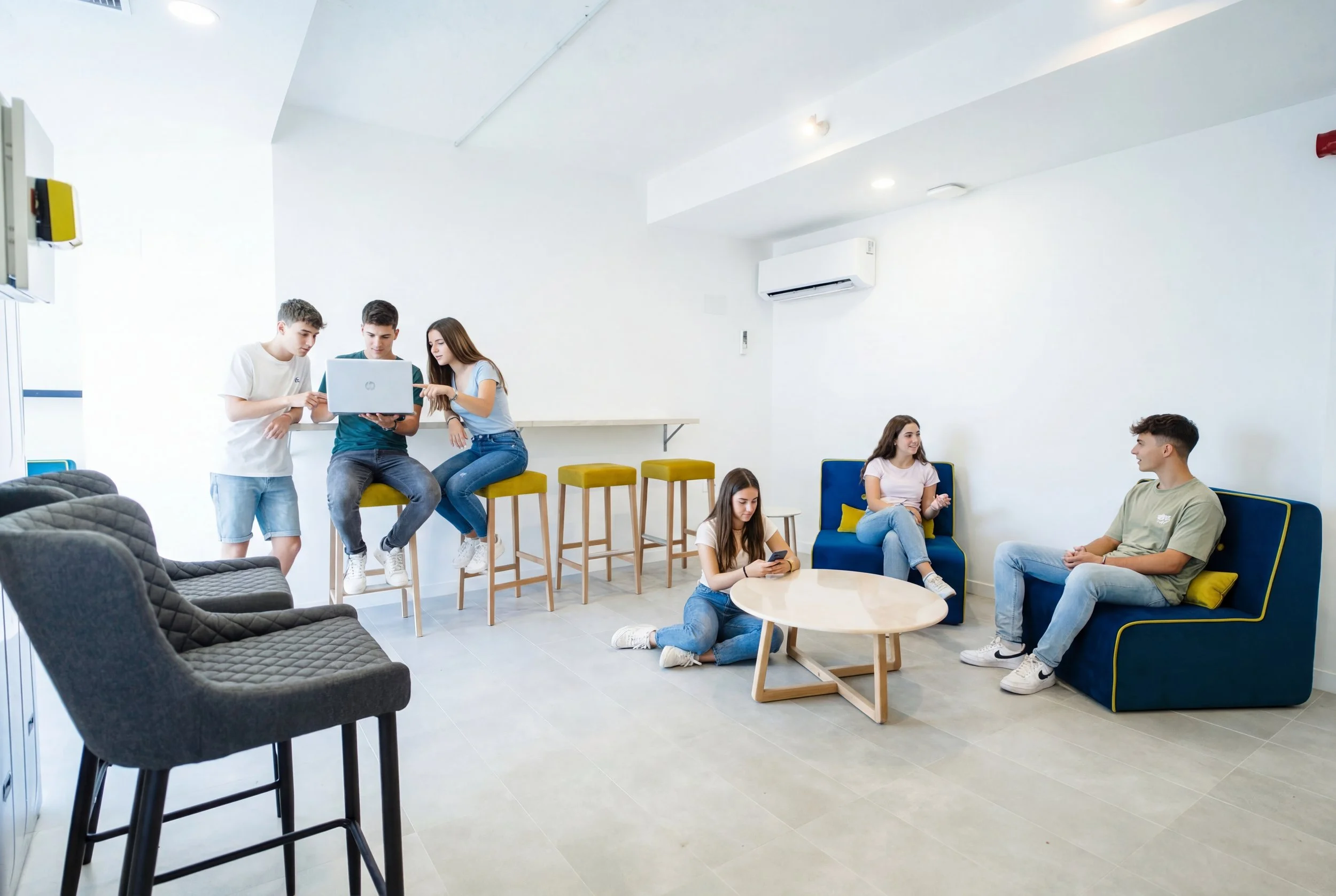 Zona común luminosa con sofás y taburetes altos; varios huéspedes charlan y usan un portátil.