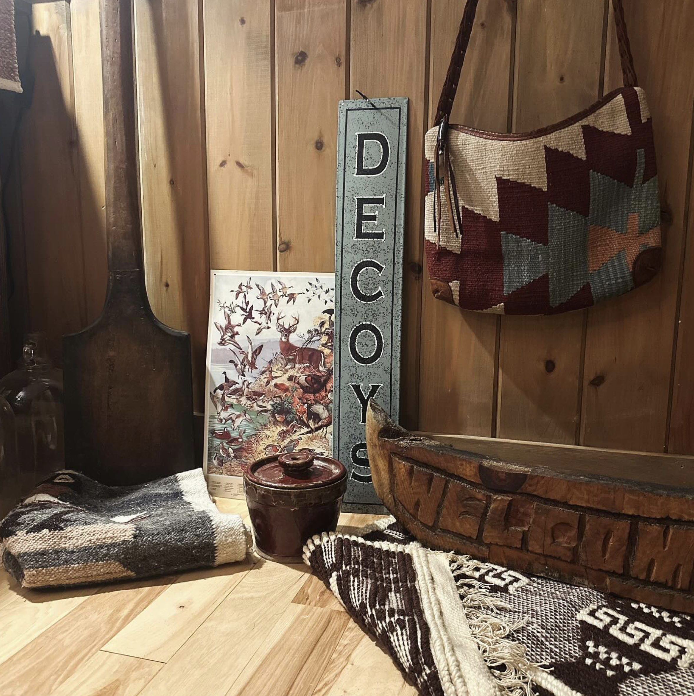 Decorative display with a wooden wall, a vintage wooden paddle, a framed artwork of birds and deer, a tall metal sign spelling "DECOY," a patterned fabric bag hanging on the wall, a ceramic jar with a lid, and rugs.