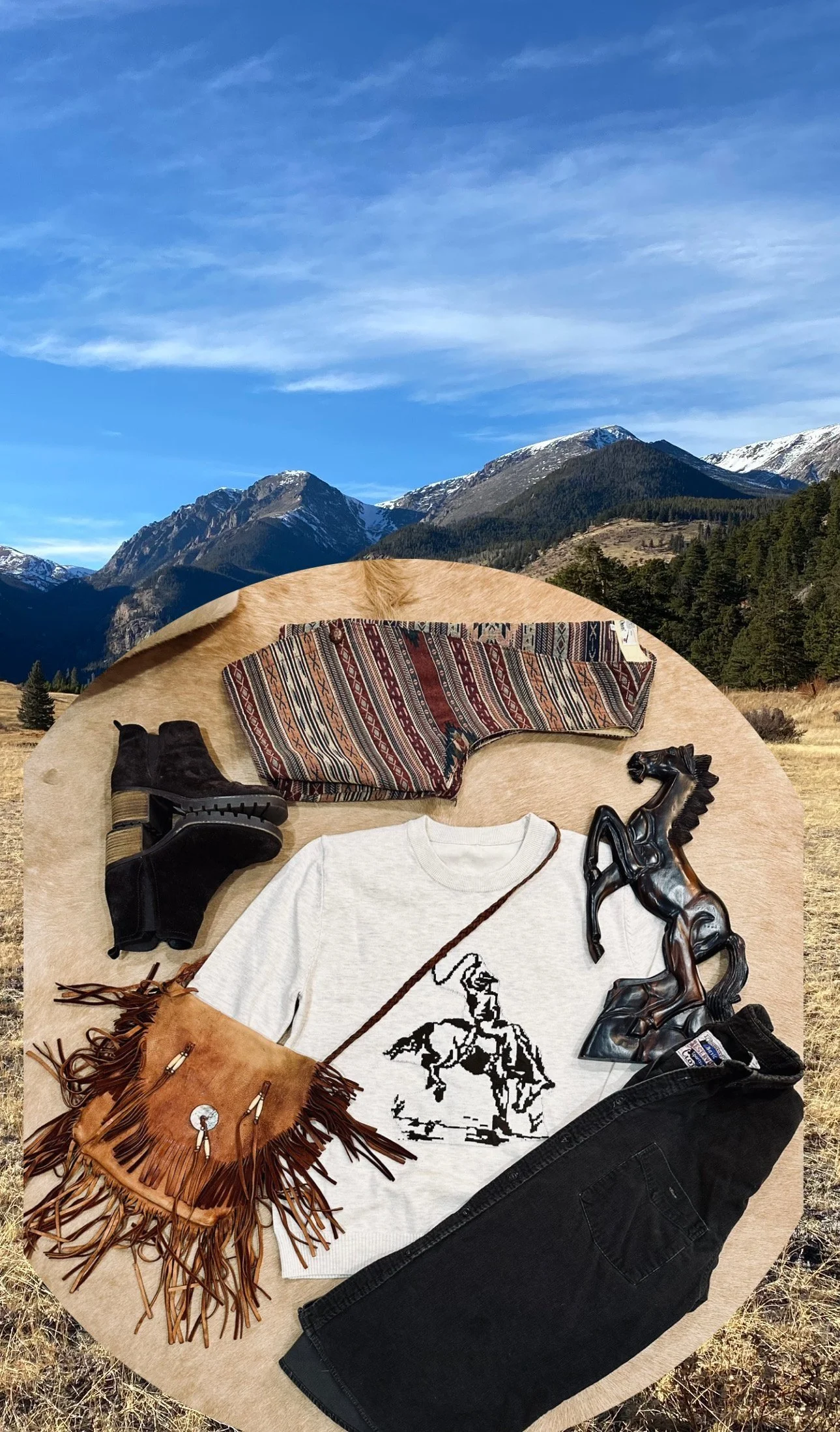 Clothing and décor items laid out on a round beige surface with a mountain and forest landscape in the background, including a patterned blanket, black boots, a white graphic t-shirt, black pants, a leather fringed purse, a small black horse sculpture, and a fringed scarf.