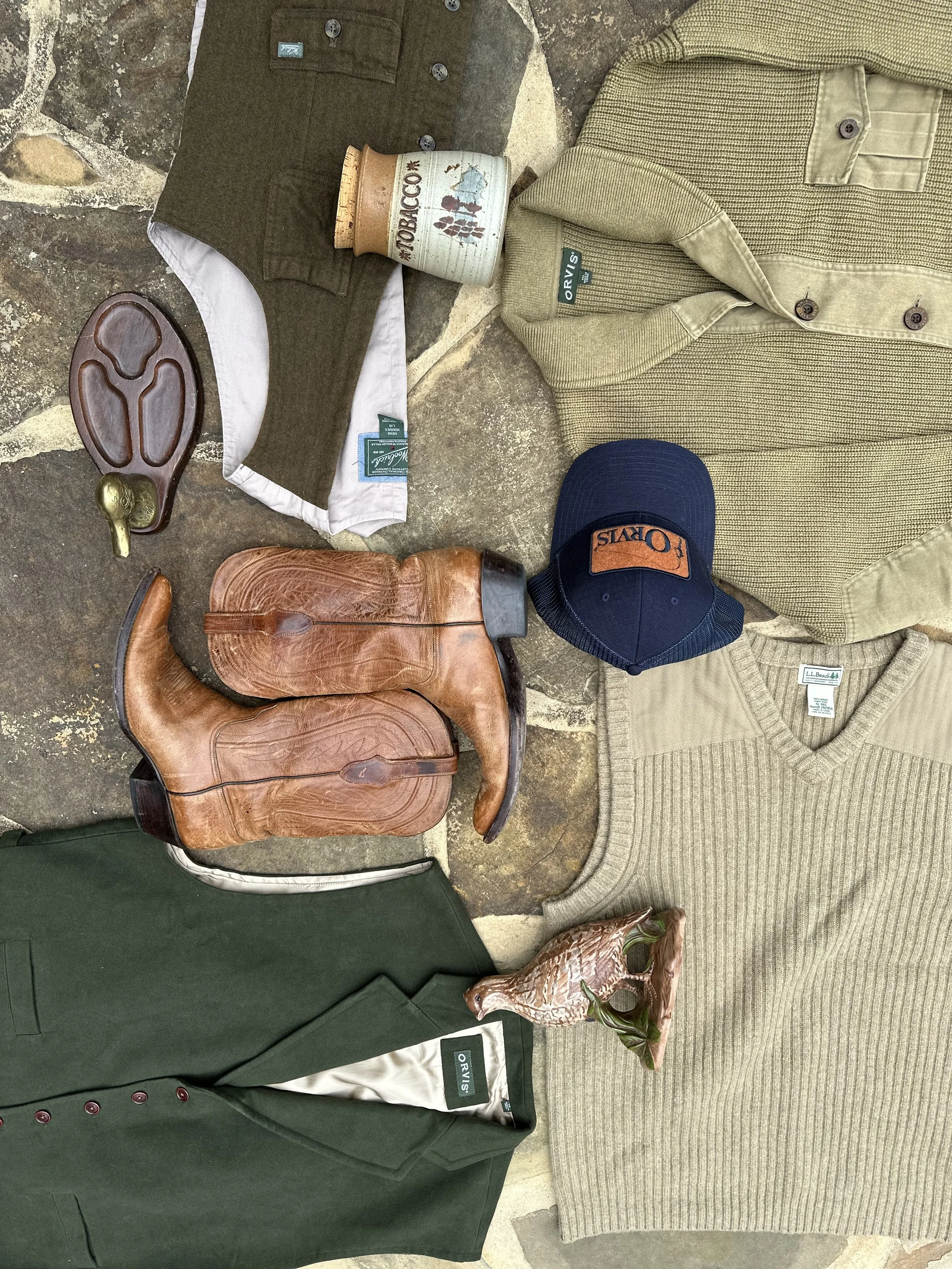 Flat lay of men's fashion and accessories, including brown cowboy boots, a green blazer, a tan sweater, a cap with "Olay" logo, a green shirt, a khaki jacket, a vintage tobacco tin, a ceramic pig figurine, a leather horned hat, and a wooden game piece, all arranged on a stone surface.