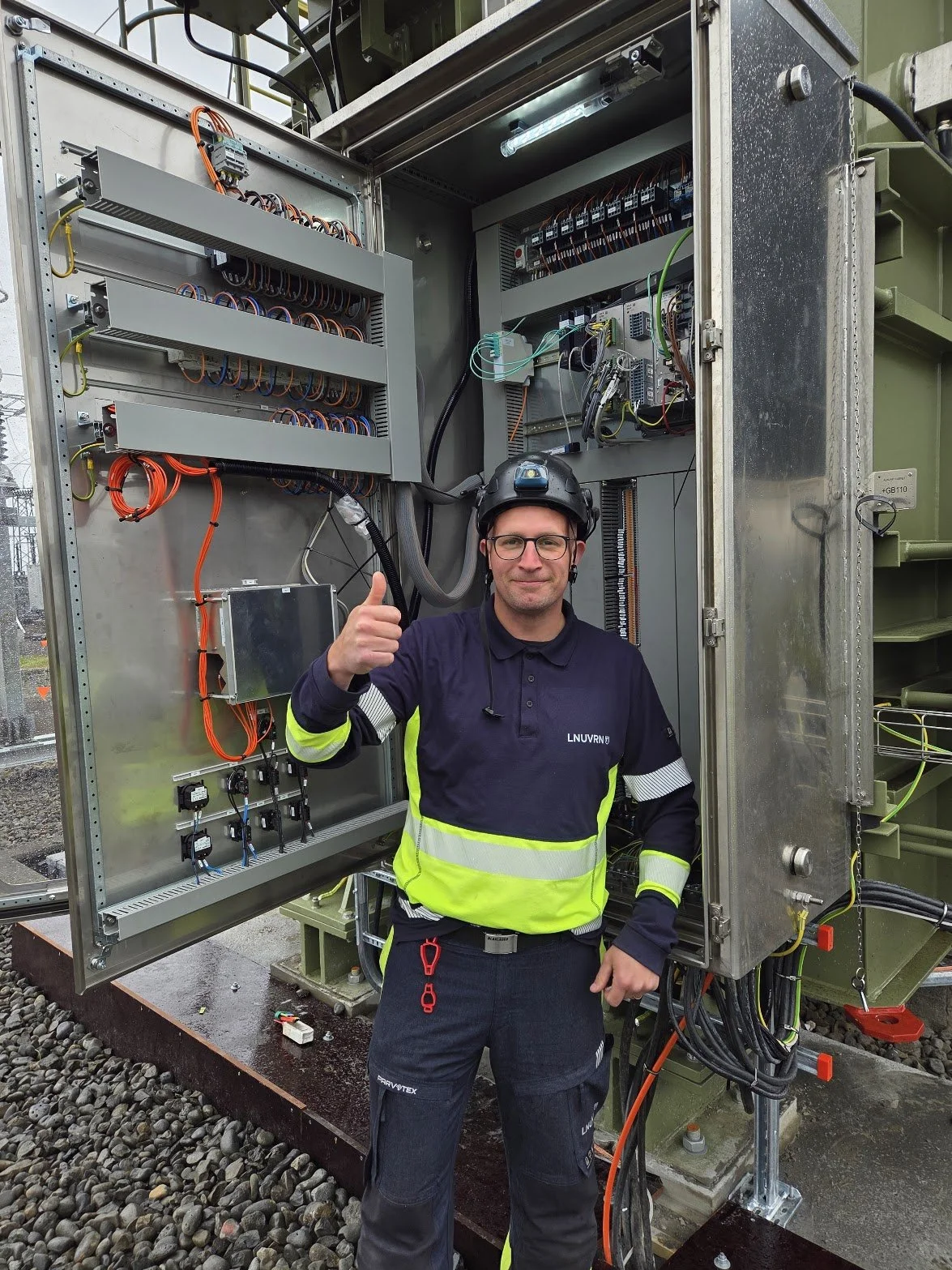 A technician wearing safety gear, including a helmet and high-visibility jacket, giving a thumbs up inside an open electrical or telecommunications cabinet with various wires and components.
