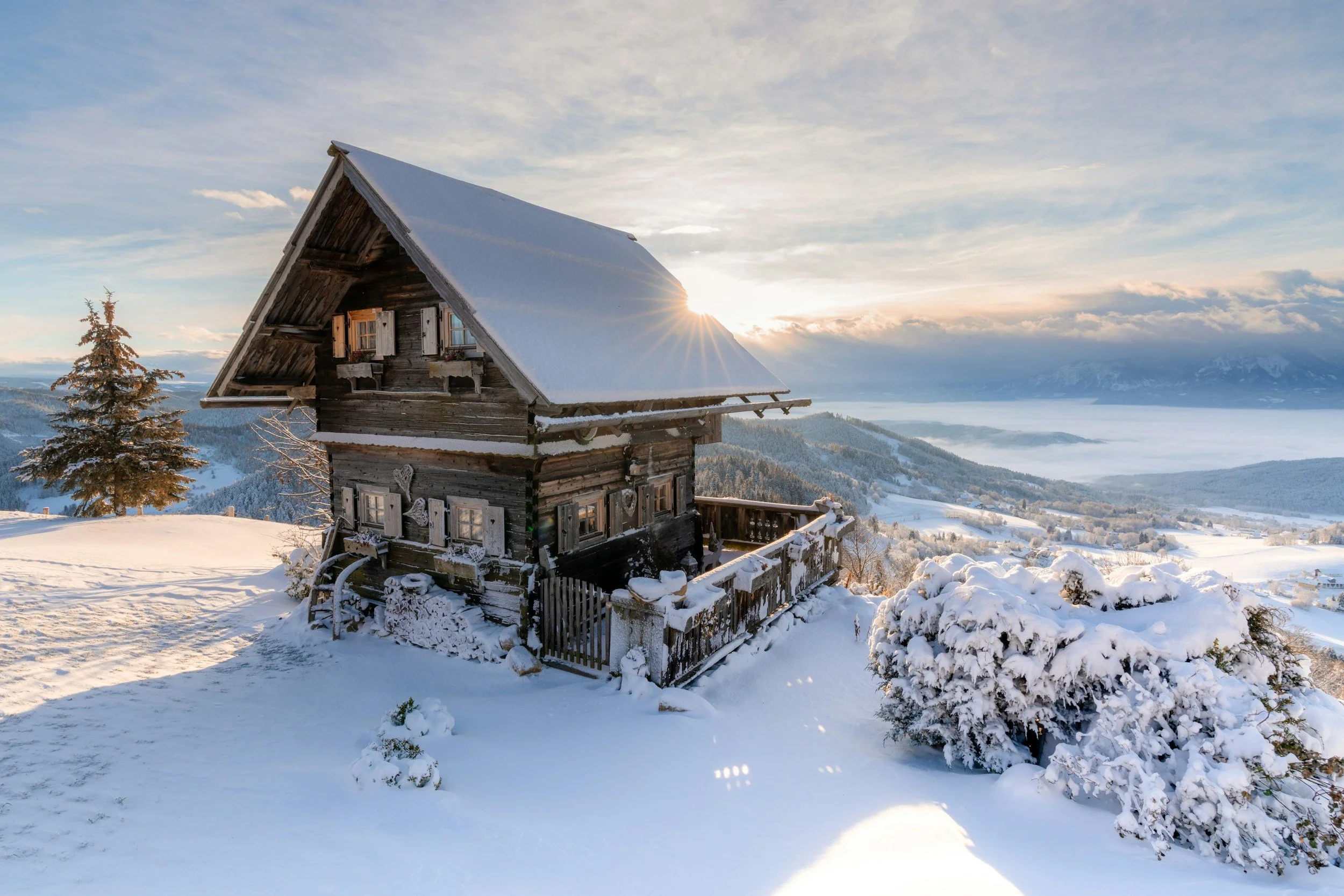 Cabane en bois recouverte de neige dans un paysage de montagne enneigée avec un ciel bleu et le soleil levant.