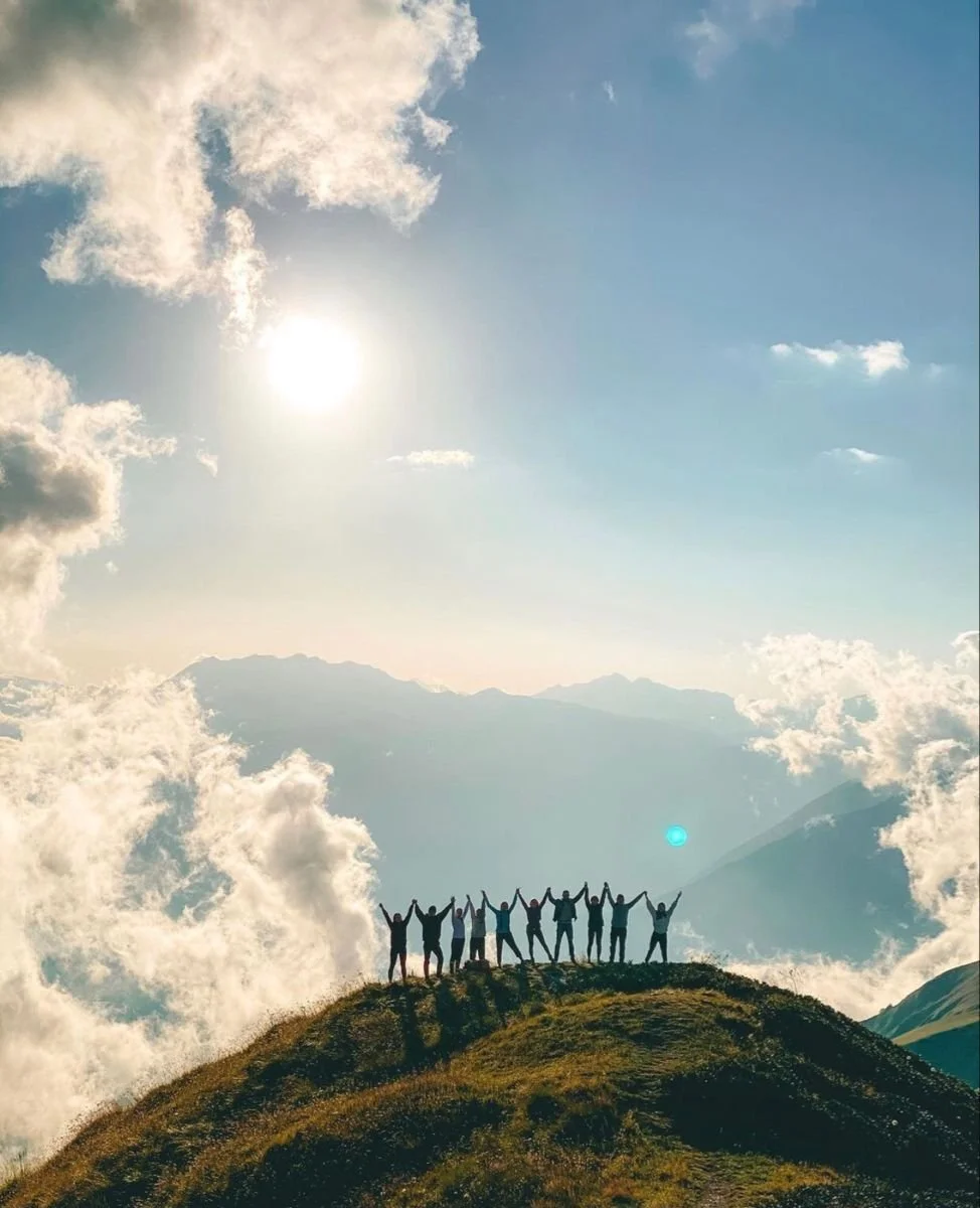 Groupe de personnes sur une colline au sommet, levant les bras en signe de victoire ou de célébration, avec un ciel ensoleillé, quelques nuages et une montagne en arrière-plan.