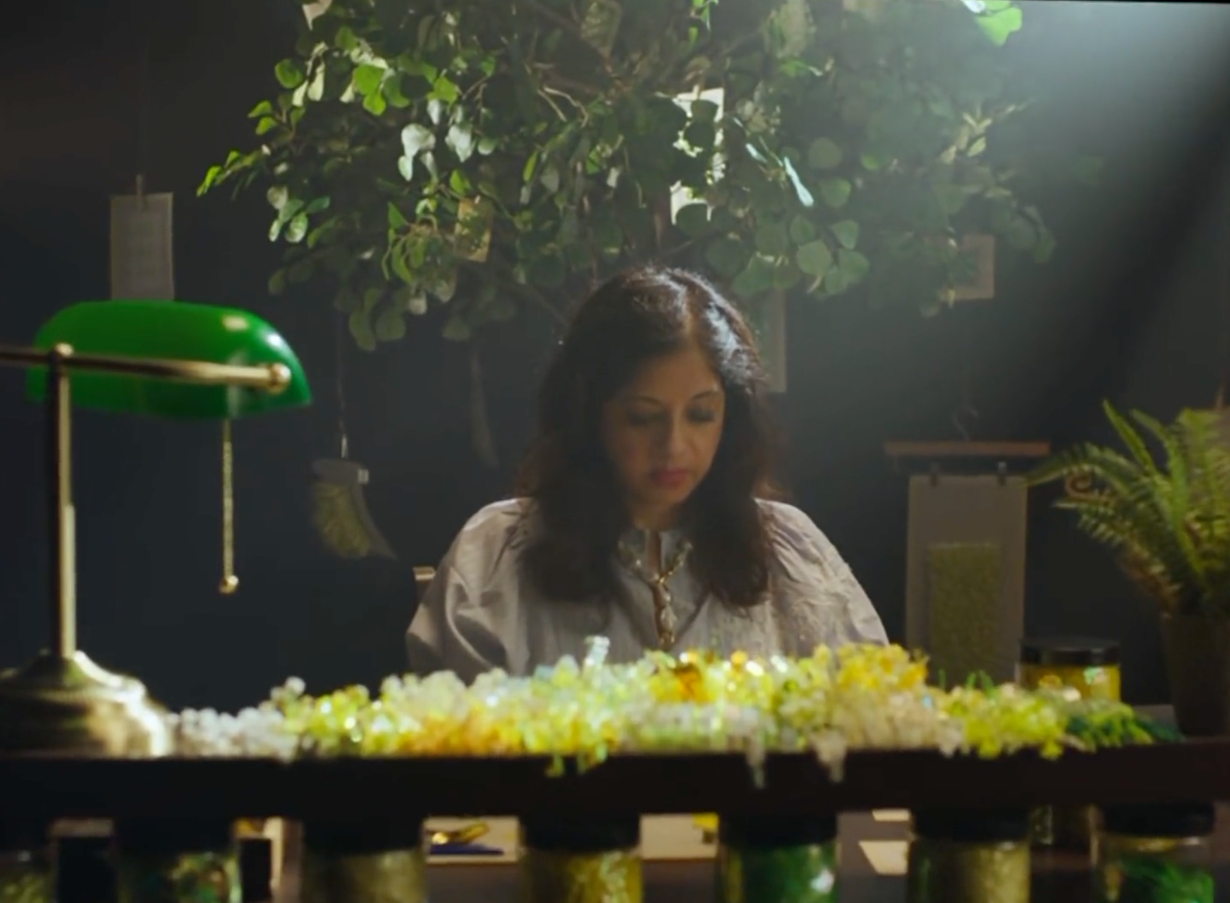 Woman sitting at her embroidery station surrounded by yellow flowers, with a large leafy plant in the background and a green banker's lamp in the foreground.