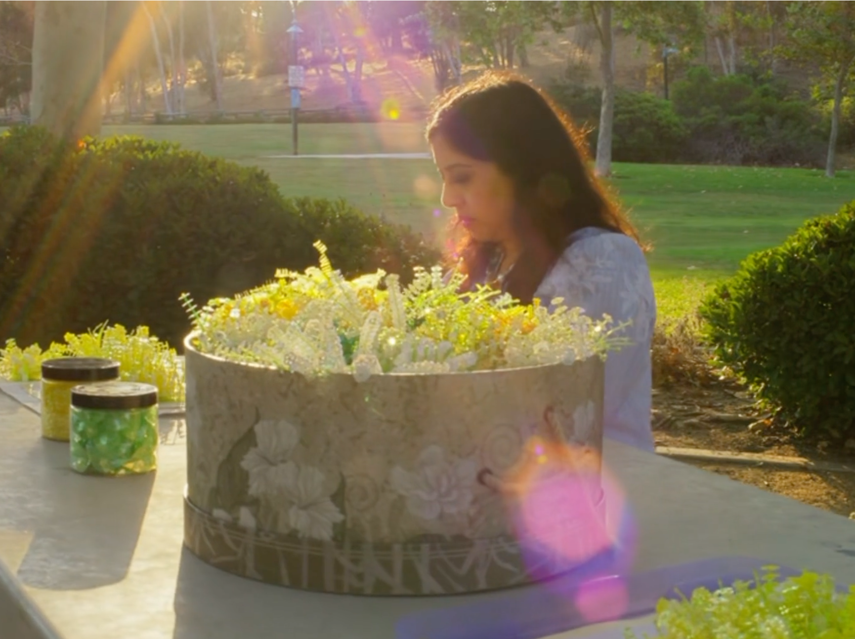 A woman with long dark hair and a white dress sitting outdoors at a table working on floral inspired embroideries
