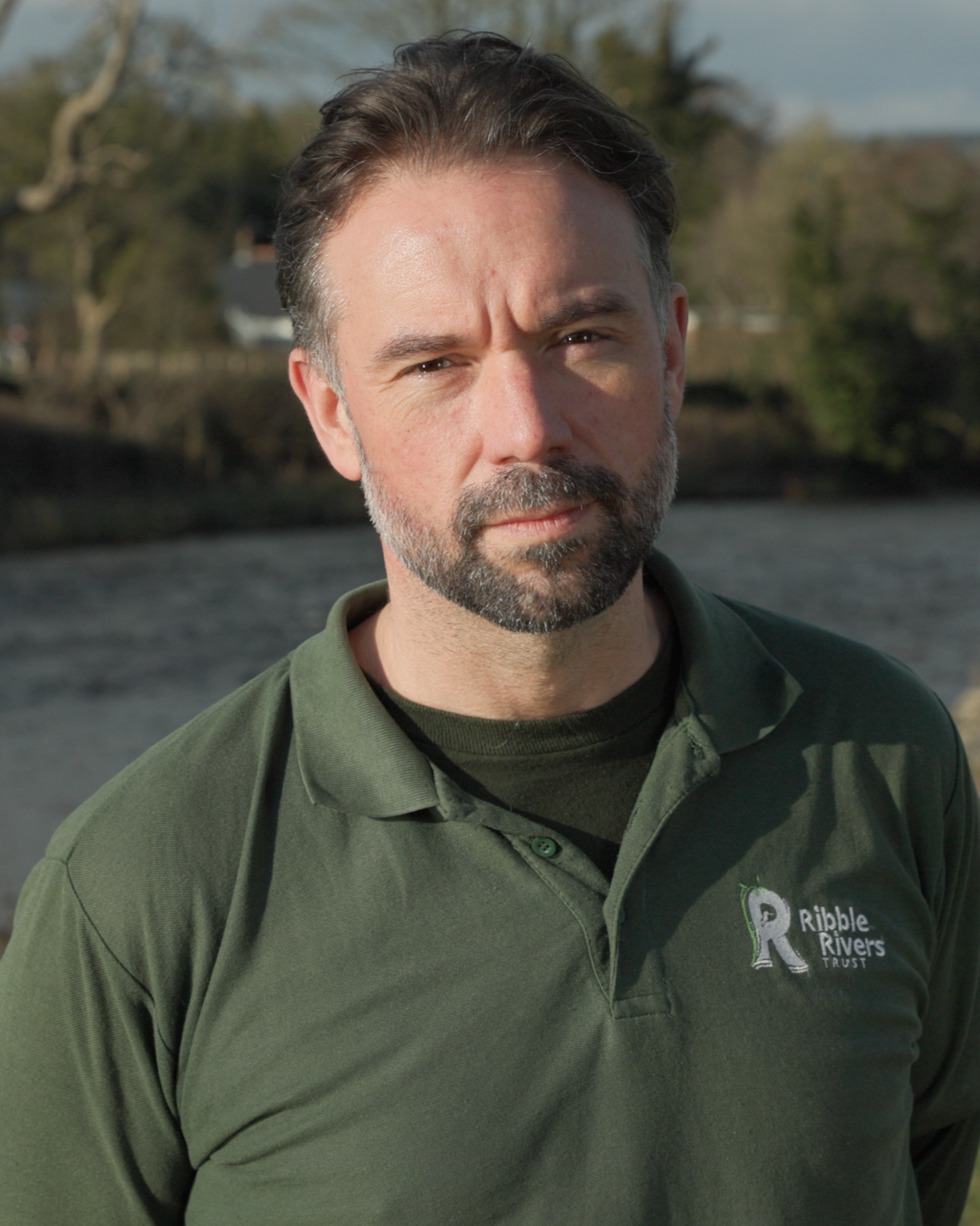 Jack Spees from Ribble Rivers Trust, standing outdoors near a body of water with trees in the background.