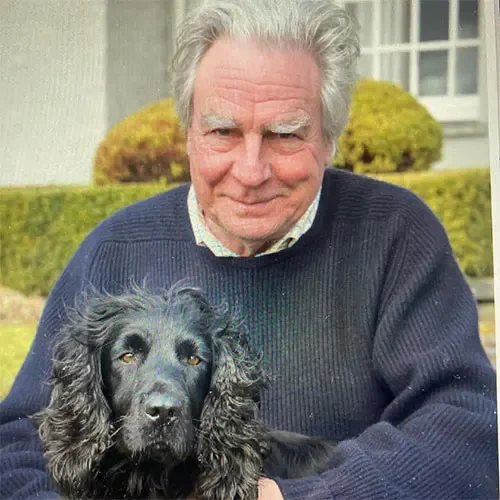 David Profumo from Country Life in a navy blue sweater smiling and holding a black Cocker Spaniel dog outdoors in front of a house with bushes.