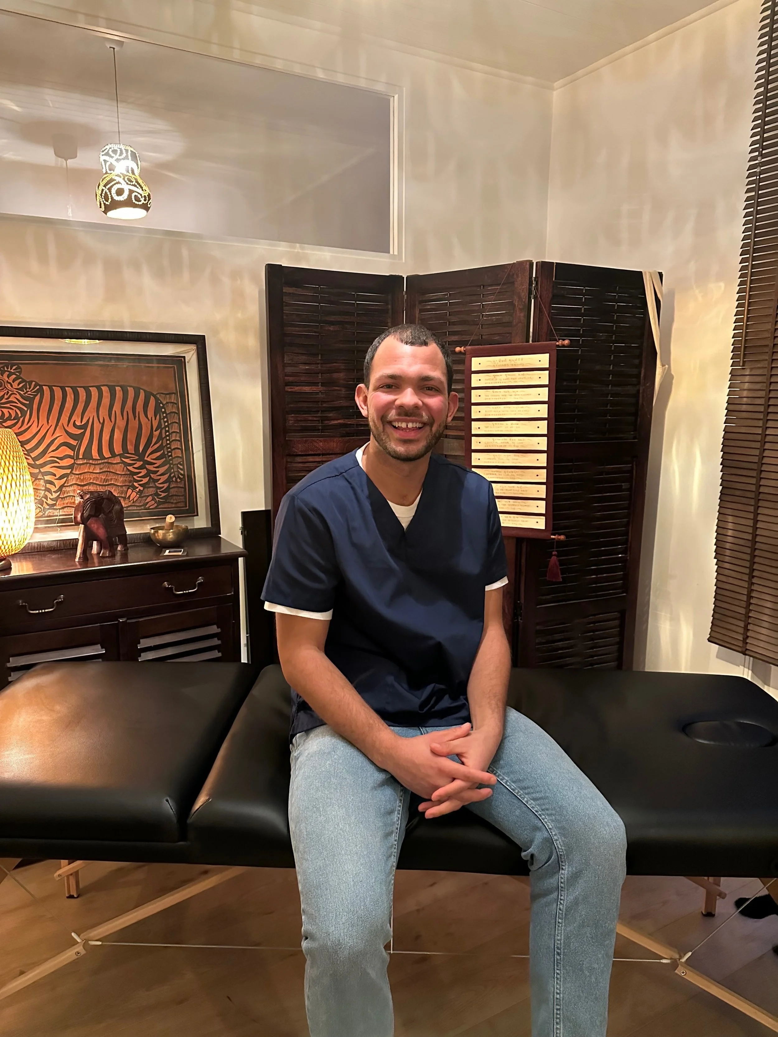 Un homme assis sur une table de massage, souriant, en tenue décontractée, dans une pièce décorée avec des éléments asiatiques, incluant une sculpture d'un éléphant et un tableau avec un tigre.