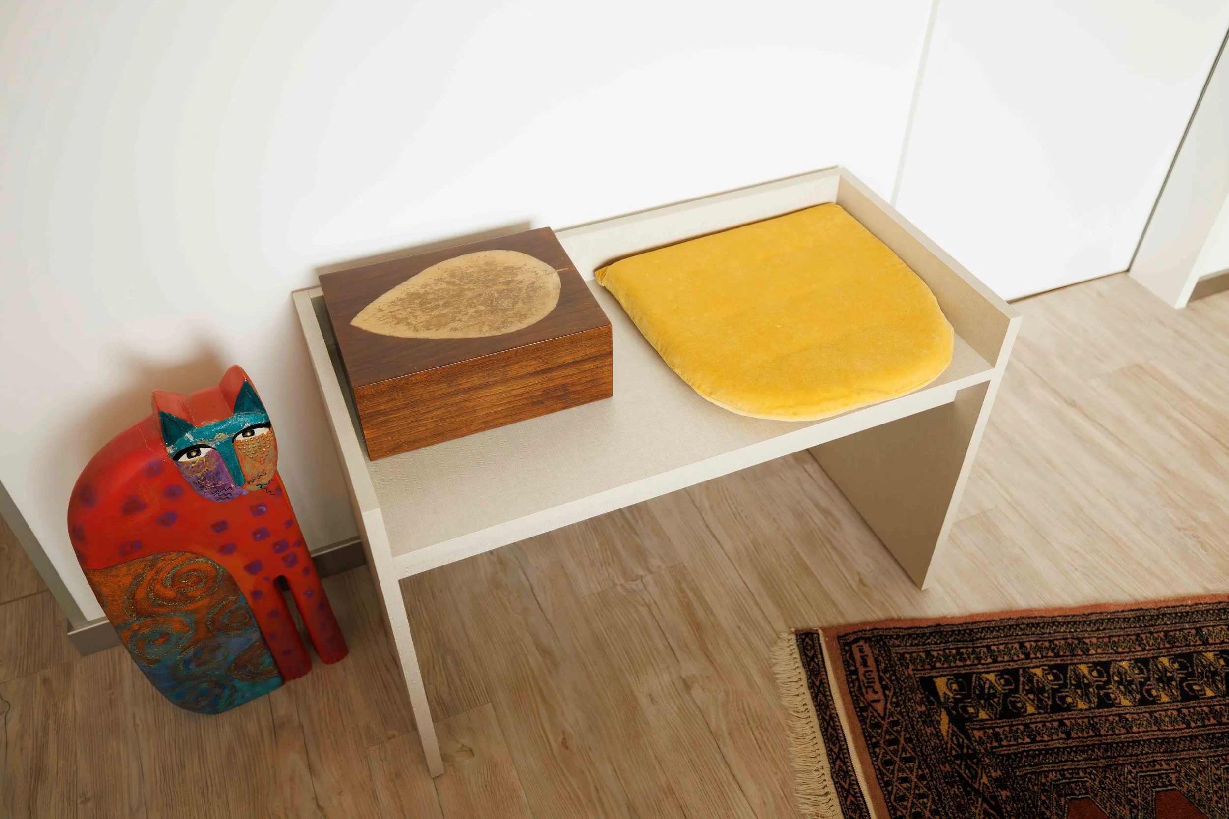 Ein weißer Konsolentisch mit einer Holzbox, einem gelben Kissen und einer roten Keramikfigur einer Katze mit buntem Gesicht, neben einem Teppich auf Holzboden.