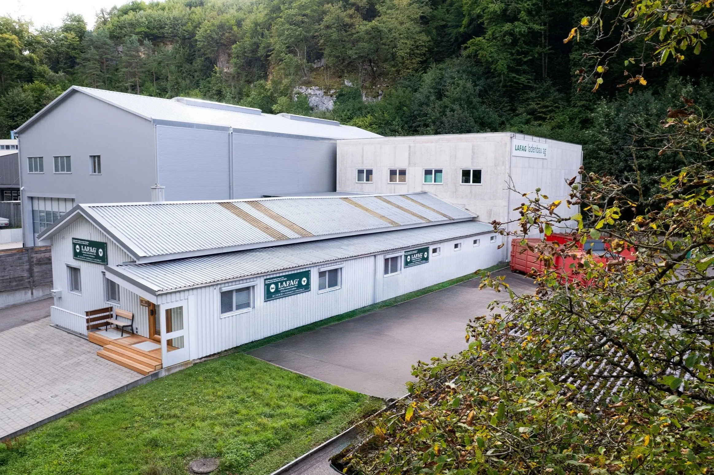 Industriegebäude mit weißen und grauen Fassaden, einige Fenster, Firmenlogos 'LAFAG' an der Frontseite, umgeben von Grünflächen, Bäumen und einem Parkplatz, im Hintergrund bewaldete Hügel.