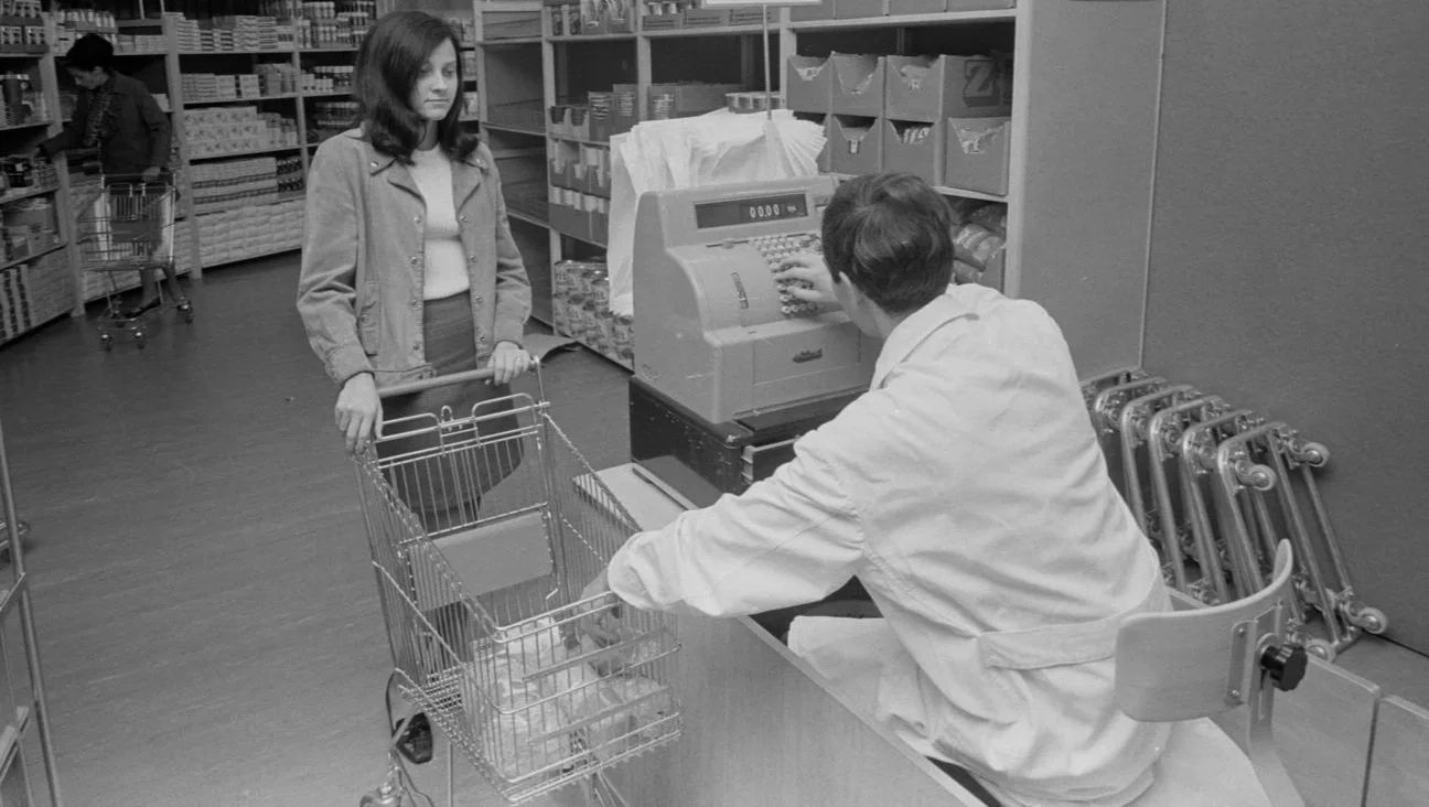 Eine junge Frau steht mit einem Einkaufswagen in einem Supermarkt vor einem Kassenautomaten, wo ein Verkäufer die Zahlung entgegennimmt.