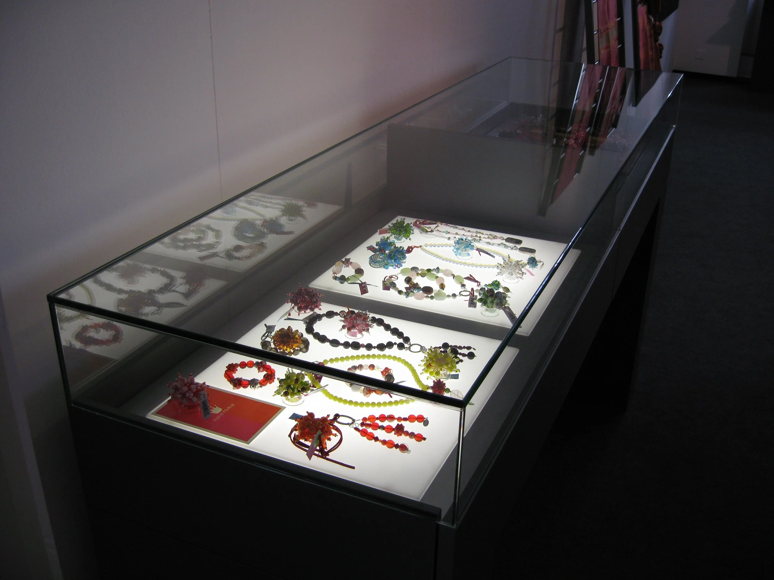 Schmuckdisplay im Museum, mit verschiedenen bunten Halsketten, Armbändern und Ohrringen in einer beleuchteten Glasvitrine
