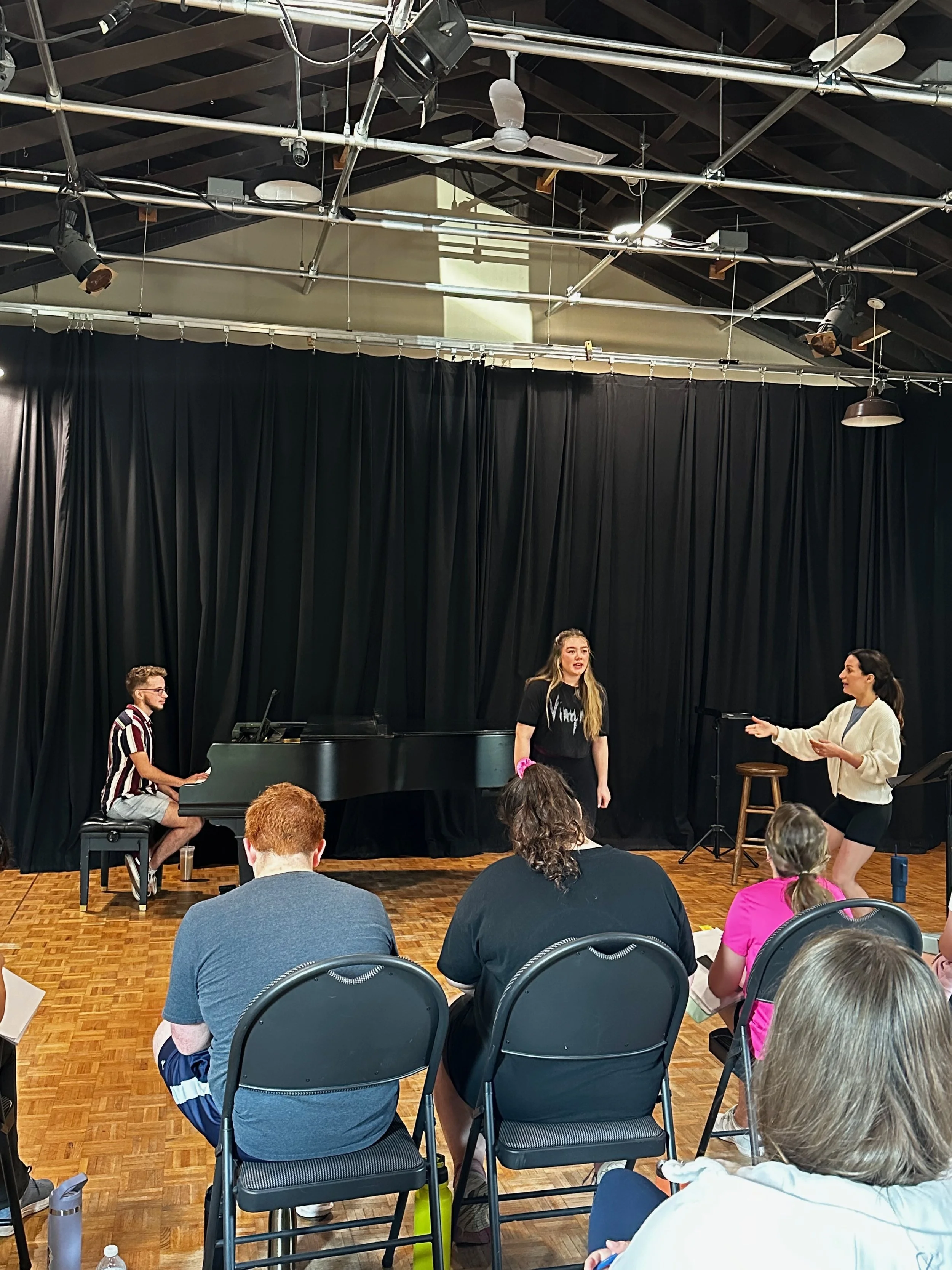 A small group of people performing and practicing in front of an audience on a stage with black curtains and a grand piano in a room with wooden floors and a high ceiling.