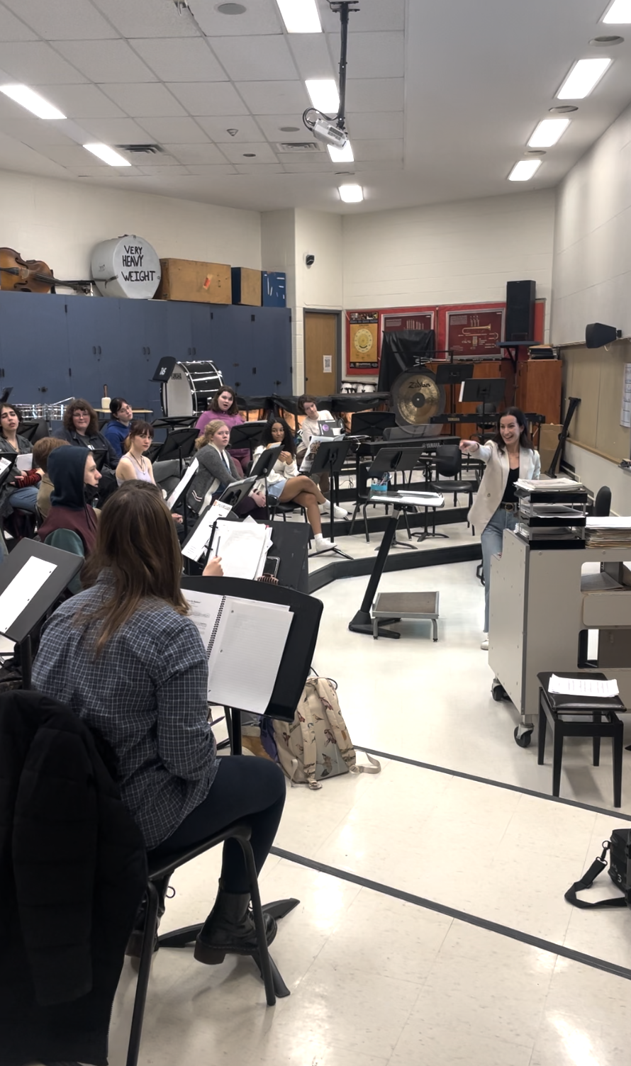 A music classroom filled with students seated with music stands, listening to a conductor or instructor leading a rehearsal, with musical instruments on shelves and a large gong in the background.