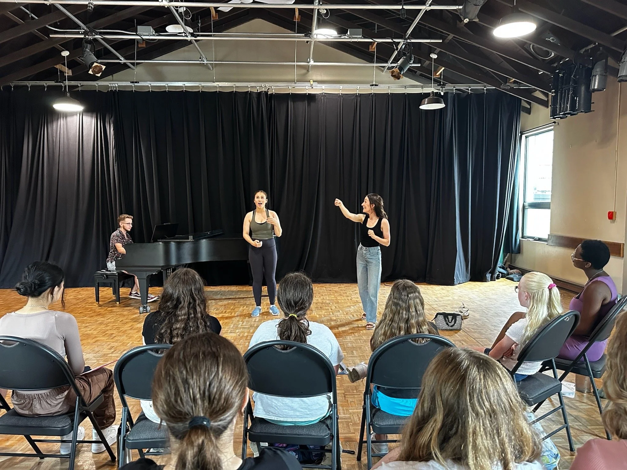A group of people watching three performers on stage in a rehearsal or show setting, with two women singing and a man playing the piano.