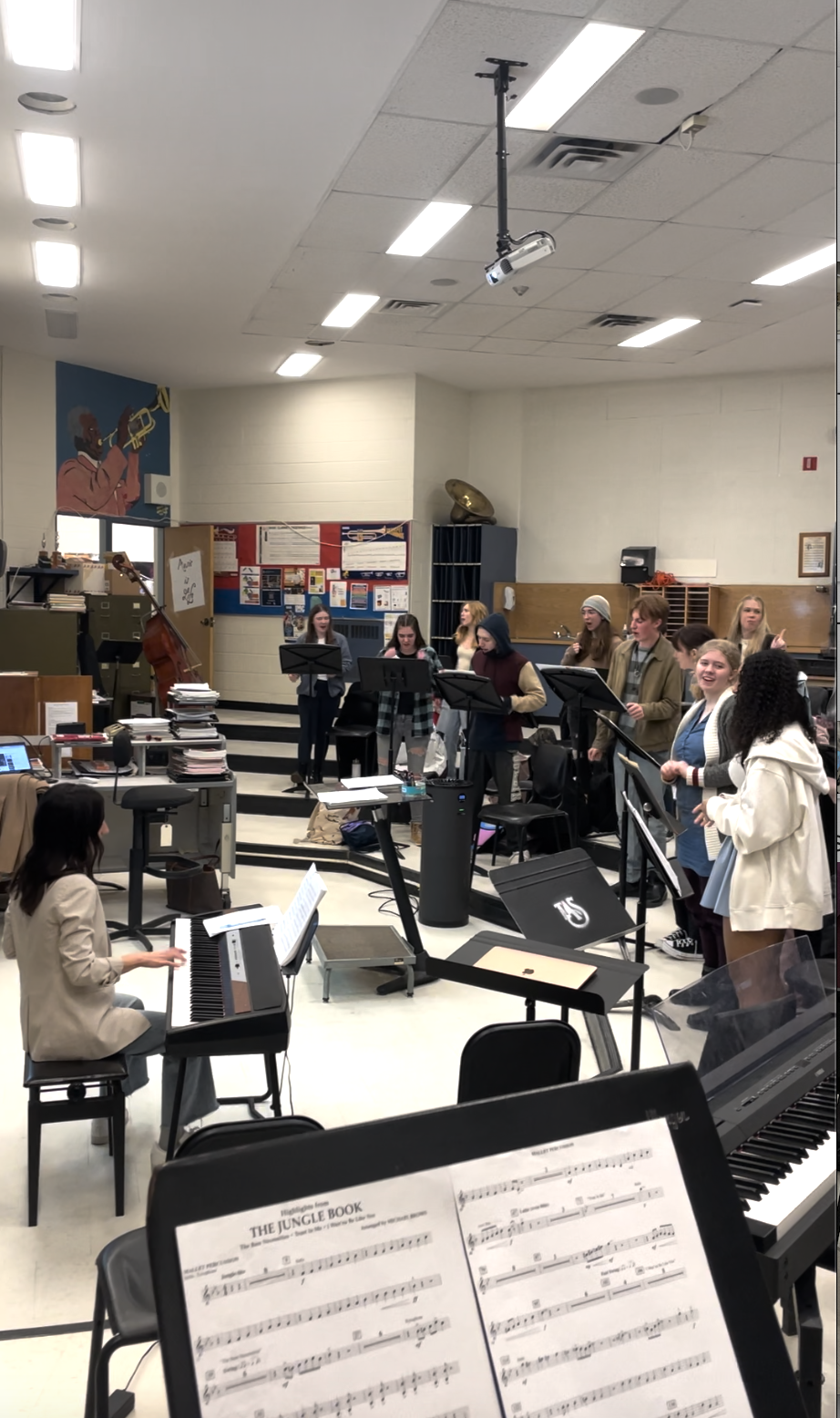 A group of students practicing choir in a music classroom, with a pianist in the foreground and music sheets on a stand.