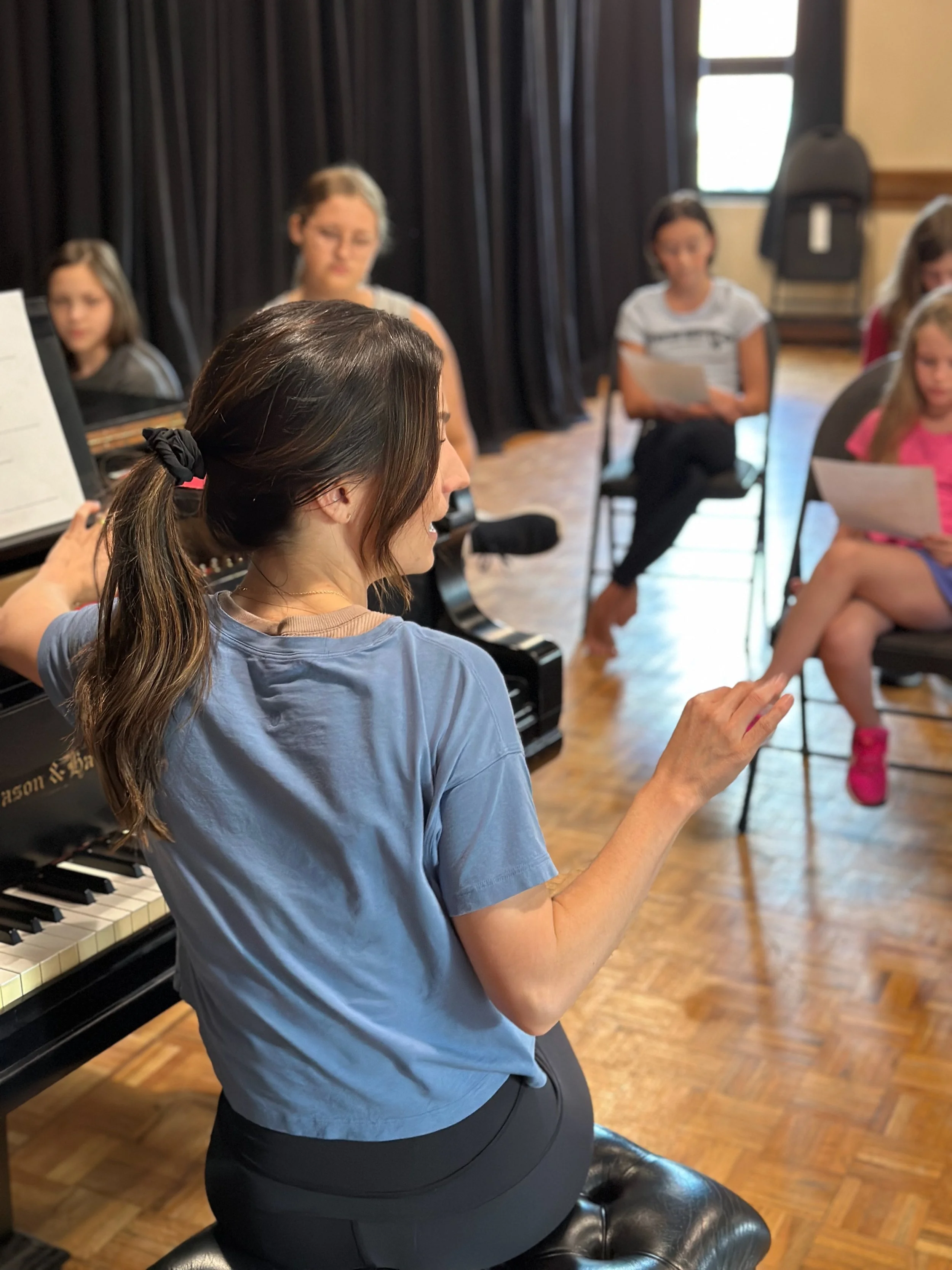 A music teacher standing near a grand piano, instructing young students seated in a semi-circle with sheet music, in a classroom with wooden floors and black curtains.