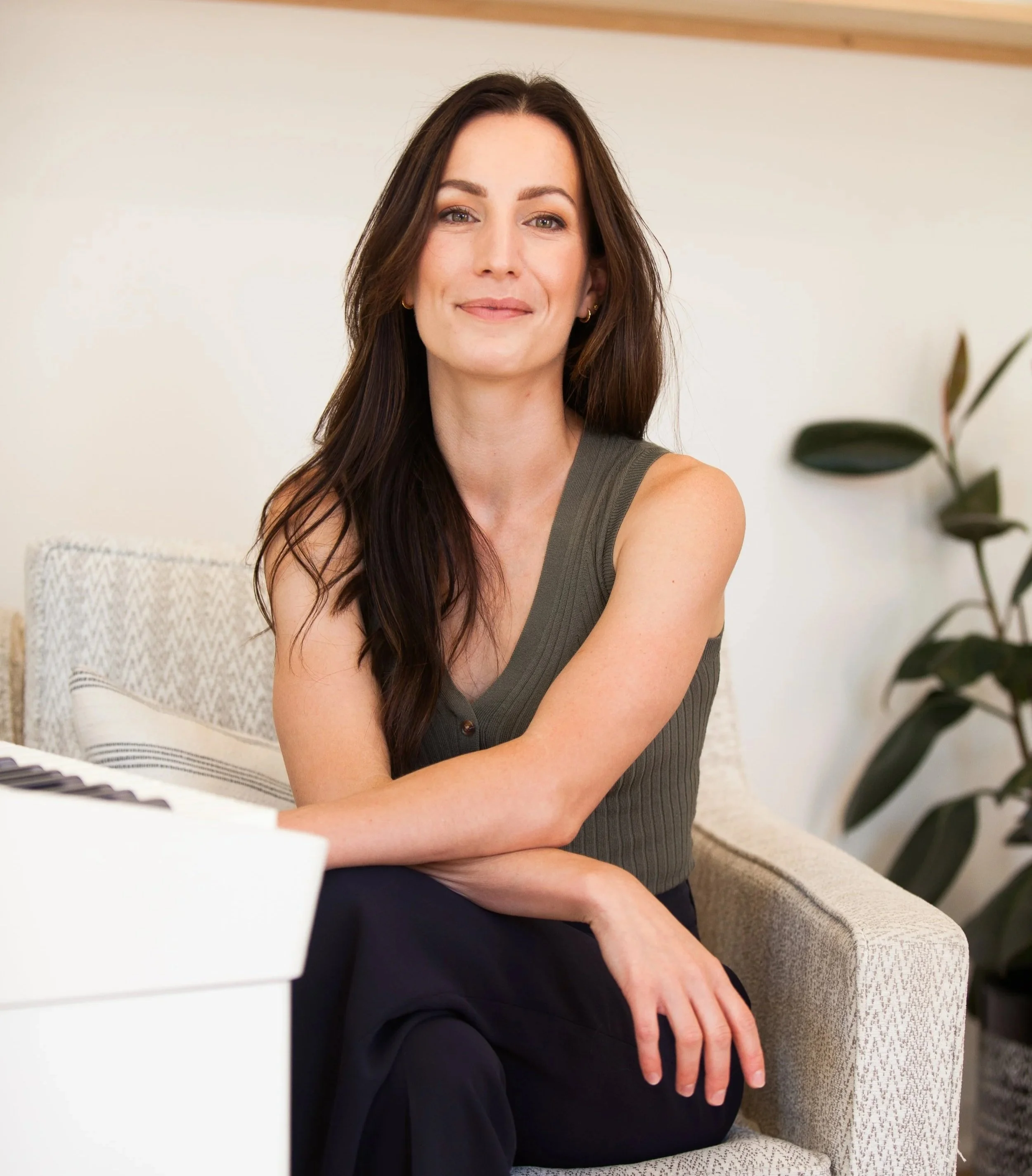 A woman with long dark hair and light skin sitting on a beige sofa next to a white keyboard, with a plant in the background.