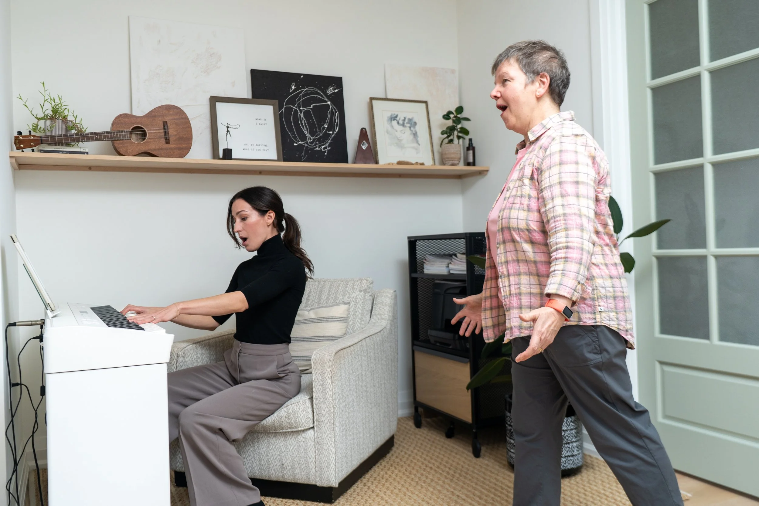 A woman giving instructions to a young girl in a music room, with a music stand and artwork on a shelf behind them.