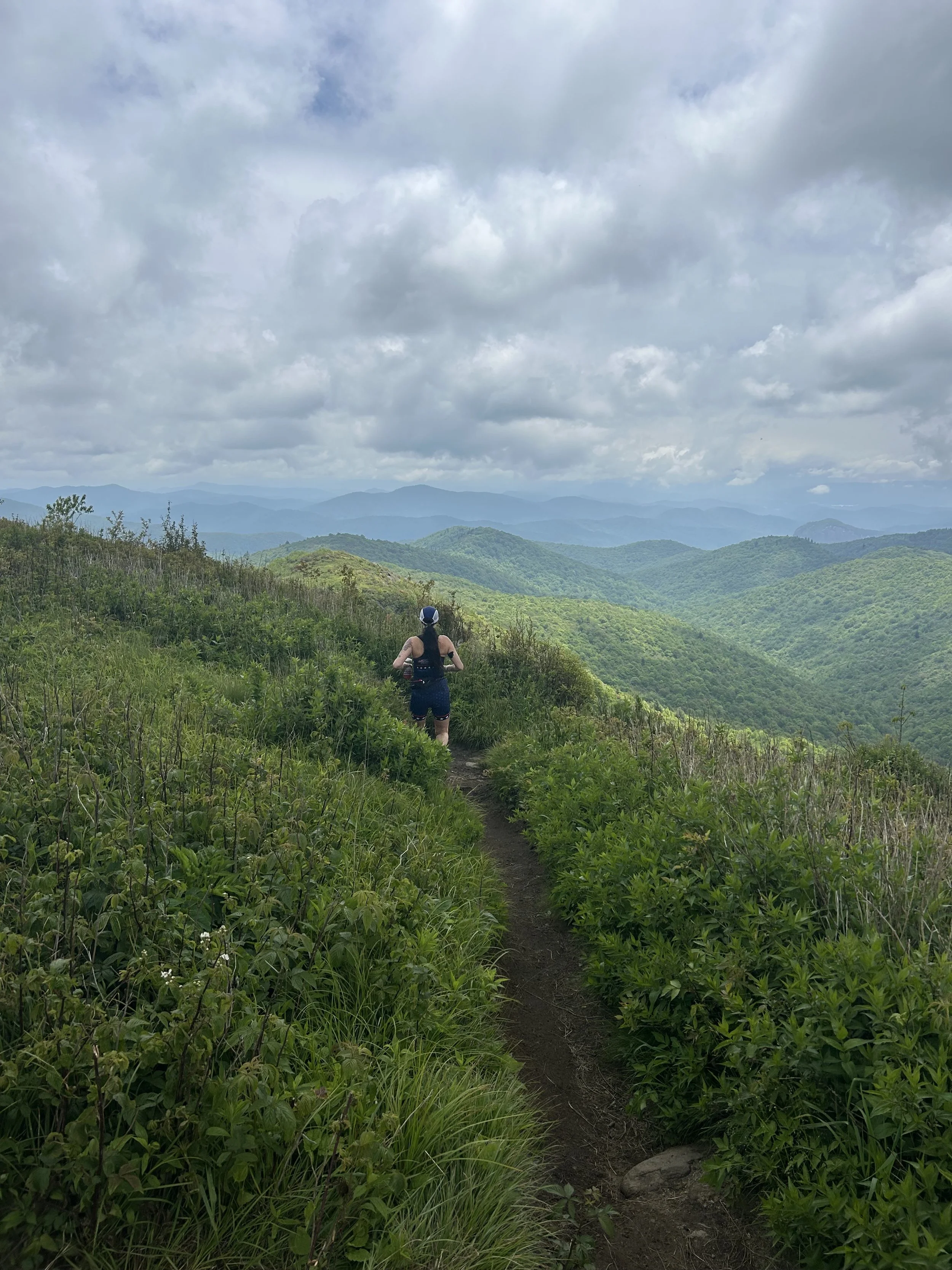 A person hiking on a narrow trail through green hills with mountain ranges and a cloudy sky in the background.