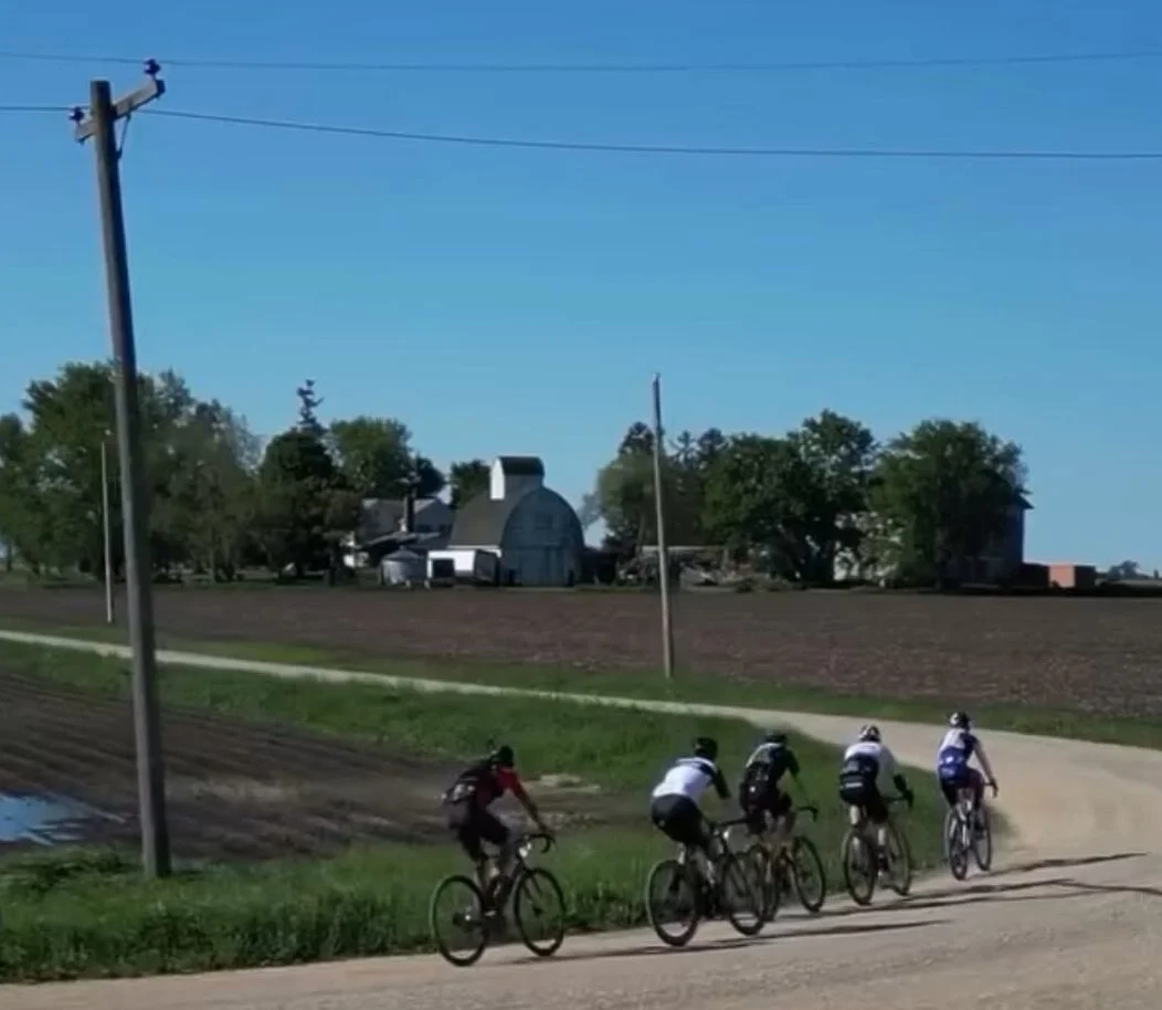 Four cyclists riding on a dirt road in a rural area with farm buildings and trees in the background under a clear blue sky.