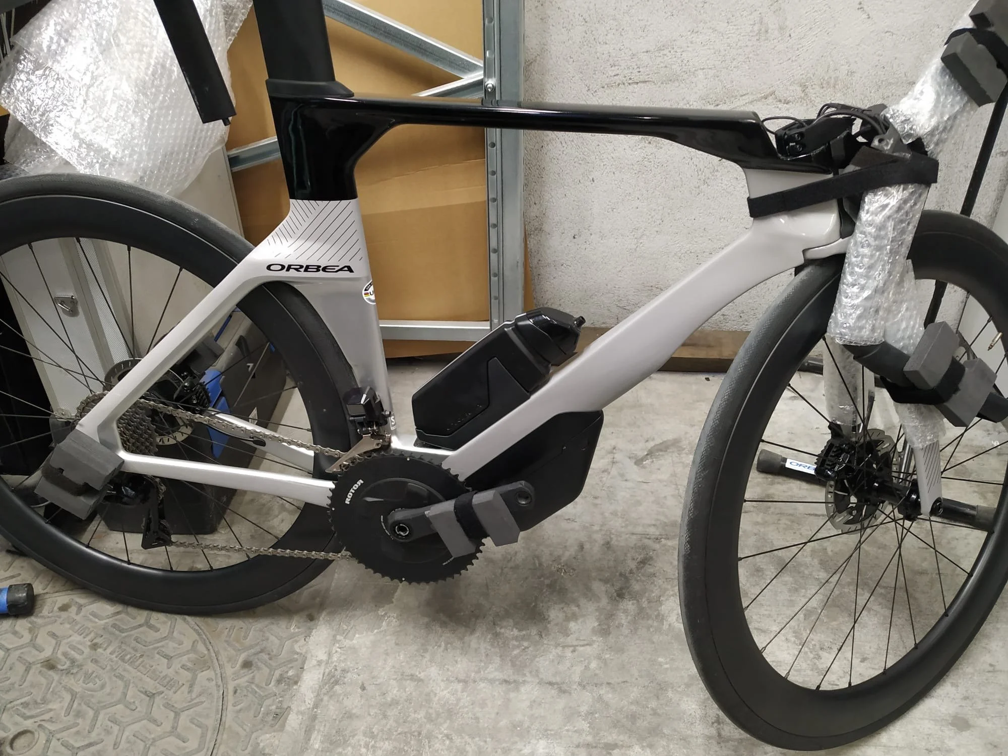 Close-up of an Orbea electric mountain bike with a black and white frame, set up indoors on a concrete floor, with a water bottle mounted on the frame.