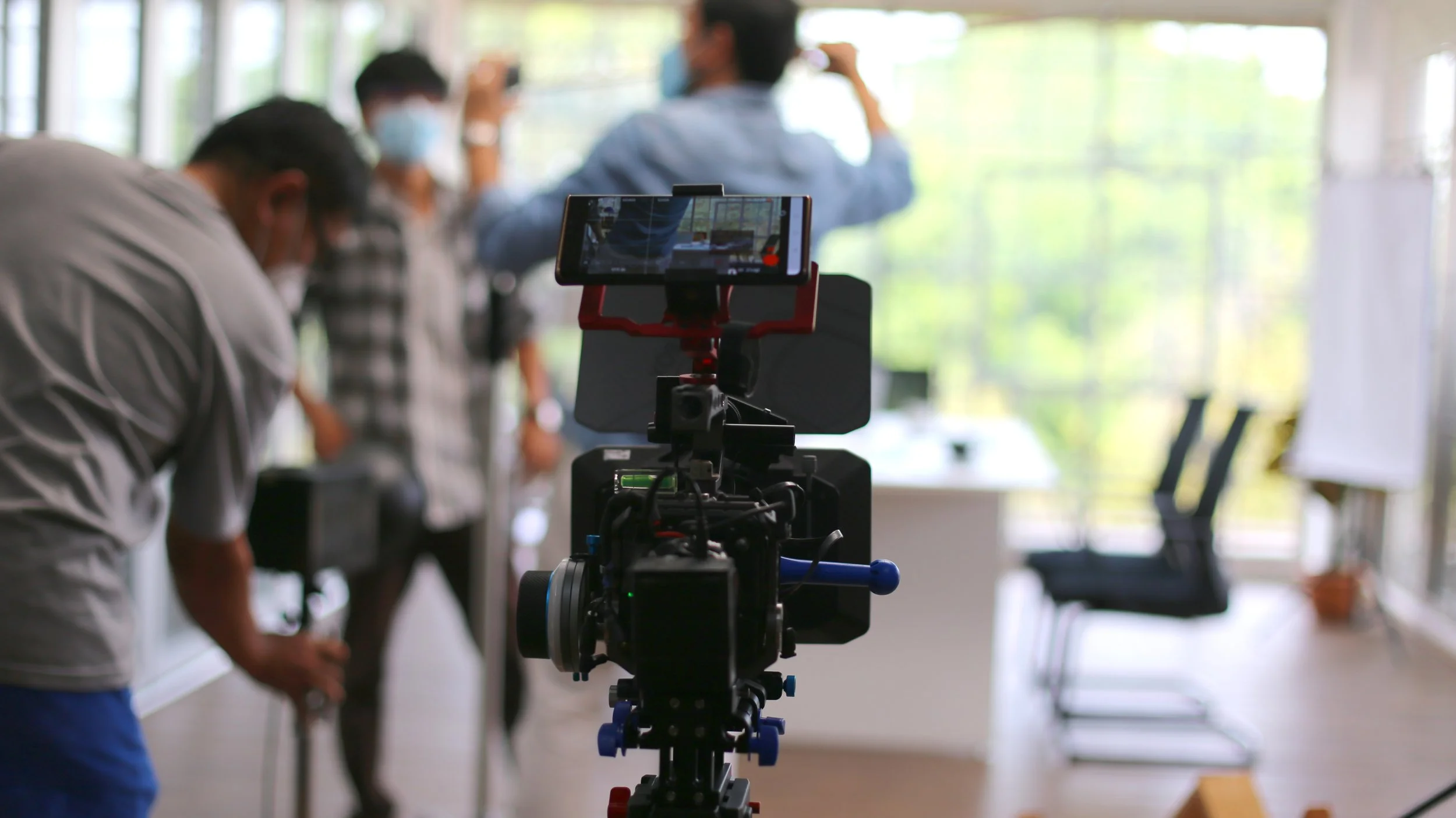 A film camera focused on a newsroom or office with several people wearing masks; one man is adjusting equipment, and others are working or recording in the background, with chairs and large windows allowing natural light.