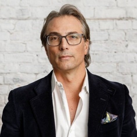 A middle-aged man with gray hair and glasses, wearing a dark blazer and white shirt, standing in front of a white brick wall.