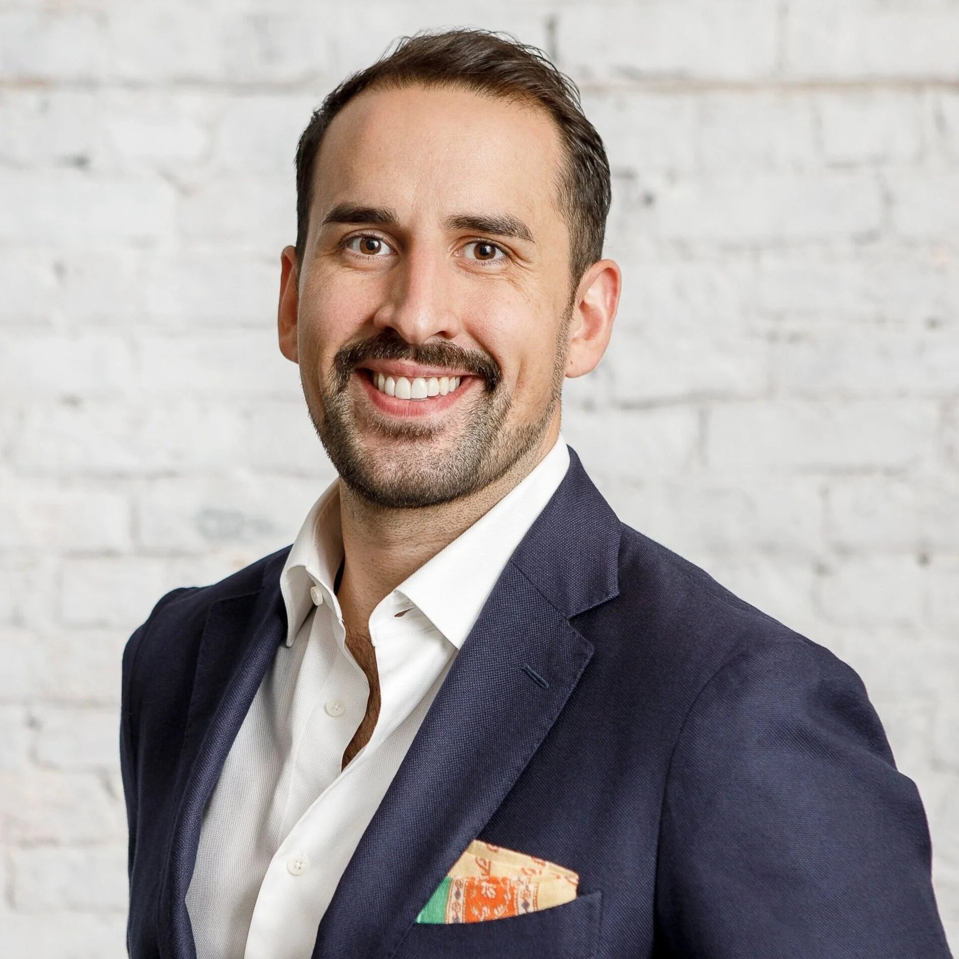 A smiling man with dark hair and a beard, wearing a navy blazer, white shirt, and a colorful pocket square, standing against a white brick wall background.