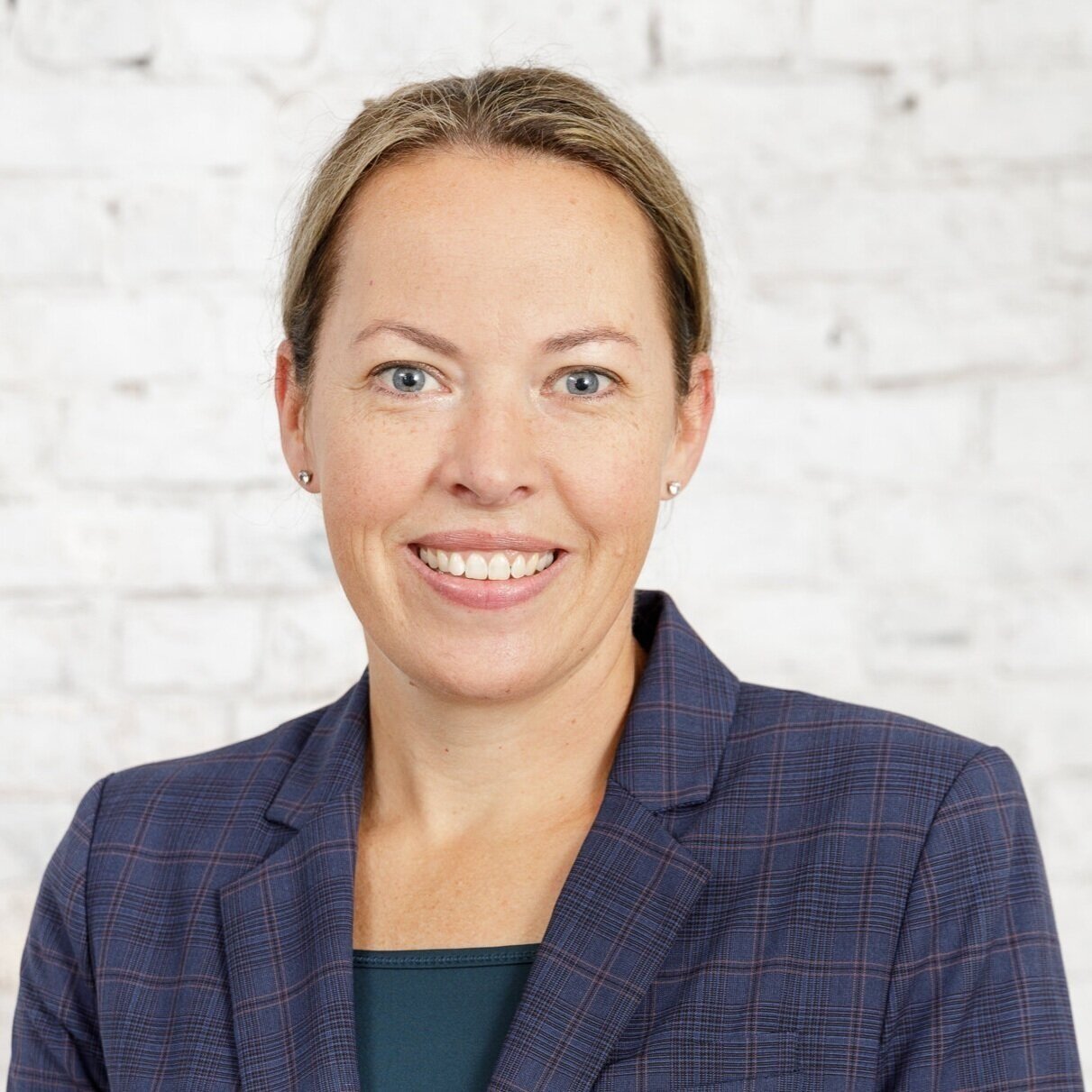 Headshot of a woman with light skin, blonde hair, and blue eyes, smiling, in front of a white brick wall, wearing a navy blue blazer.
