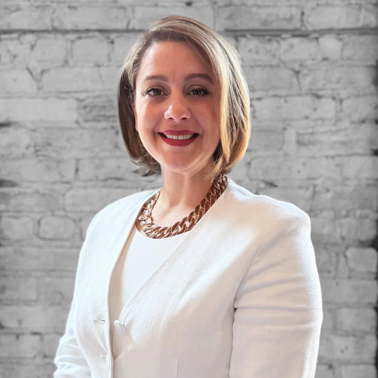 A woman with short, light brown hair wearing a white blazer, a chunky gold necklace, and smiling in front of a gray brick wall.