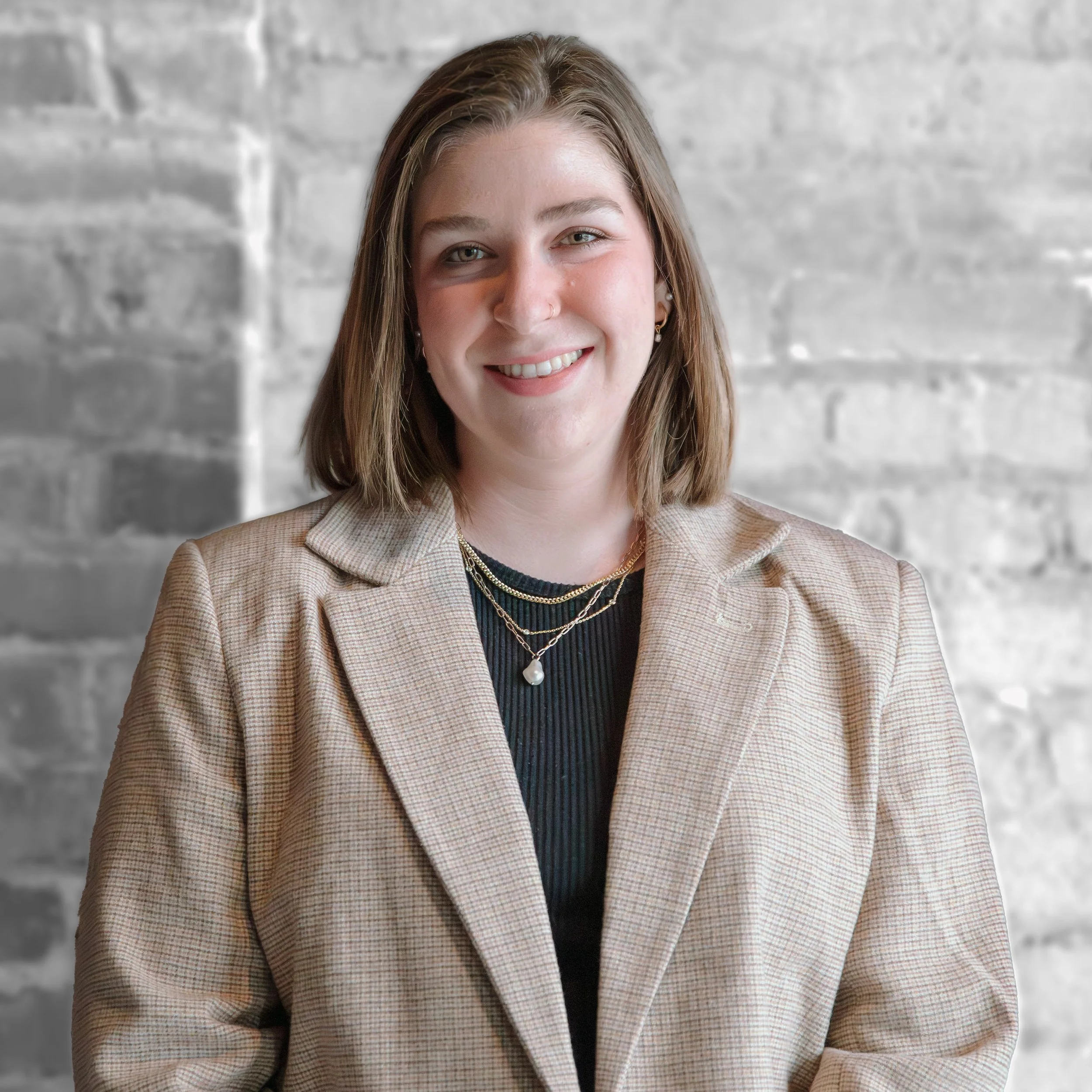 A young woman with shoulder-length brown hair and fair skin smiling, wearing a beige plaid blazer and layered necklaces, standing against a gray brick wall background.