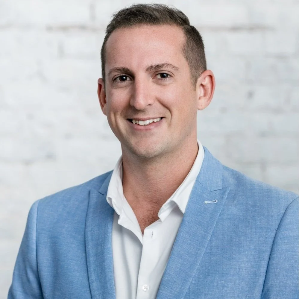Portrait of a smiling man in a blue blazer and white shirt standing in front of a white brick wall.