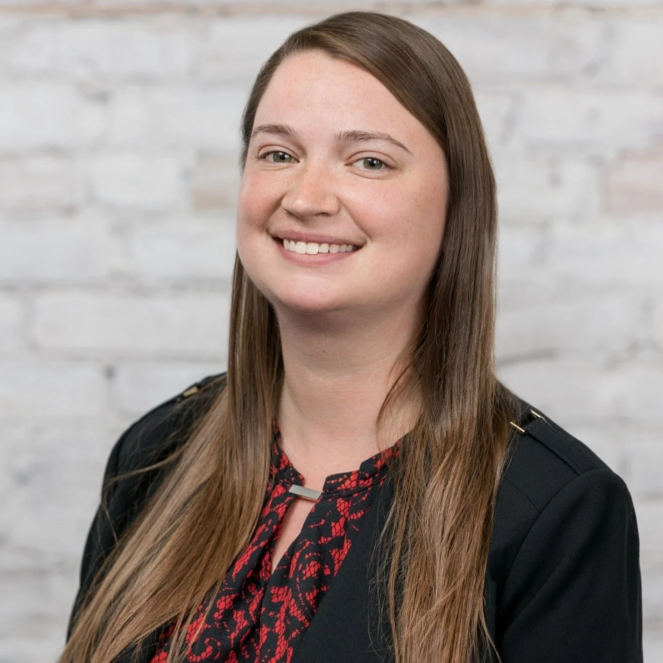 A professional woman with long brown hair, smiling, wearing a black blazer with a red and black patterned blouse, standing in front of a white brick wall.