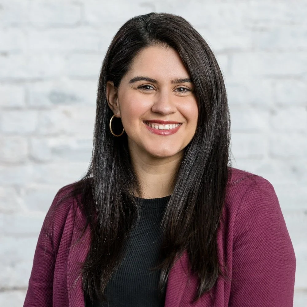 A woman with long dark hair, hoop earrings, wearing a black top and a maroon blazer, smiling in front of a light gray brick wall.